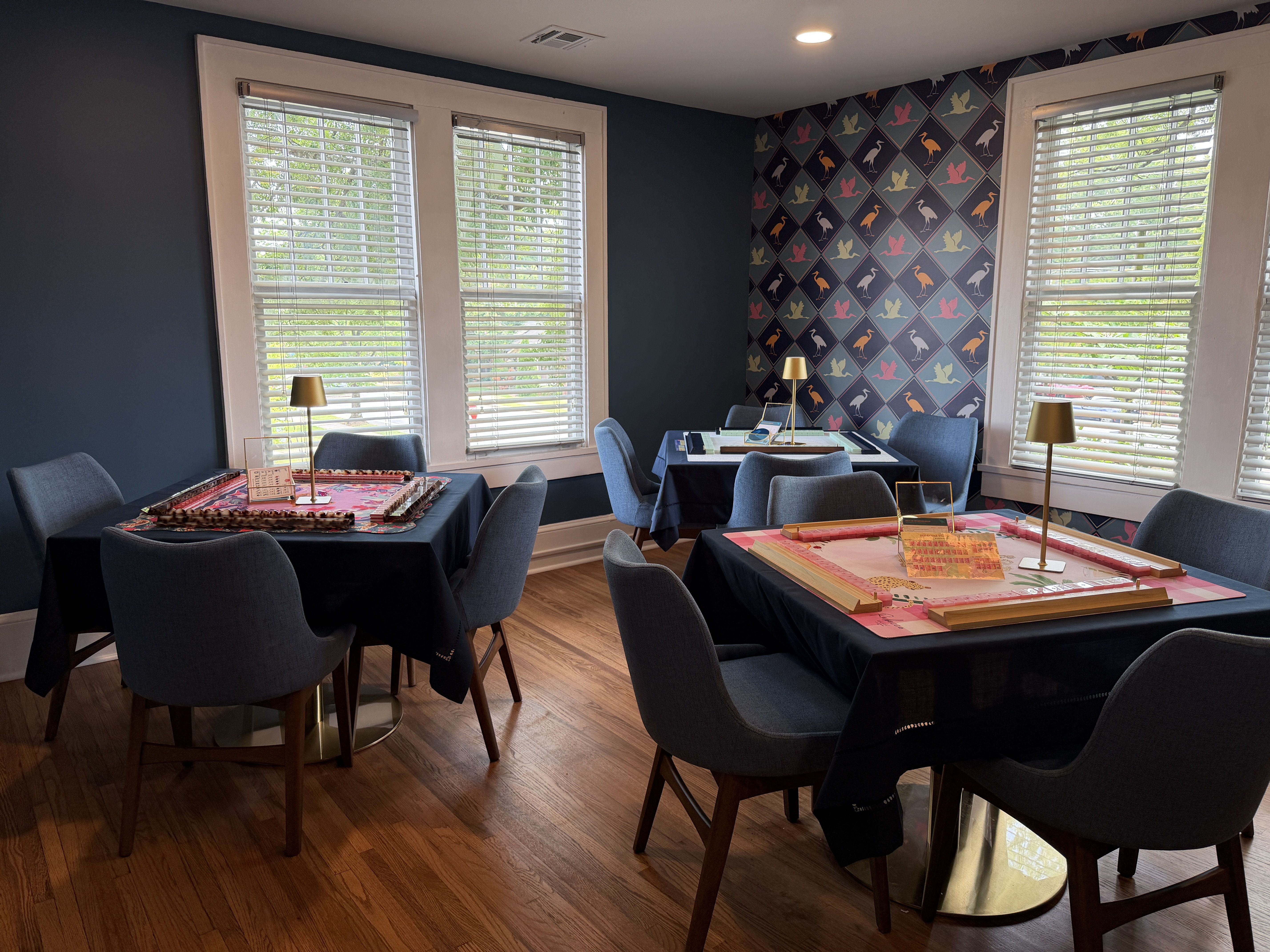 Cozy dining room with dark blue walls and a colorful diamond-pattern wallpaper featuring bird silhouettes. Tables with navy cloths, gray chairs, gold lamps, and large windows with white blinds.