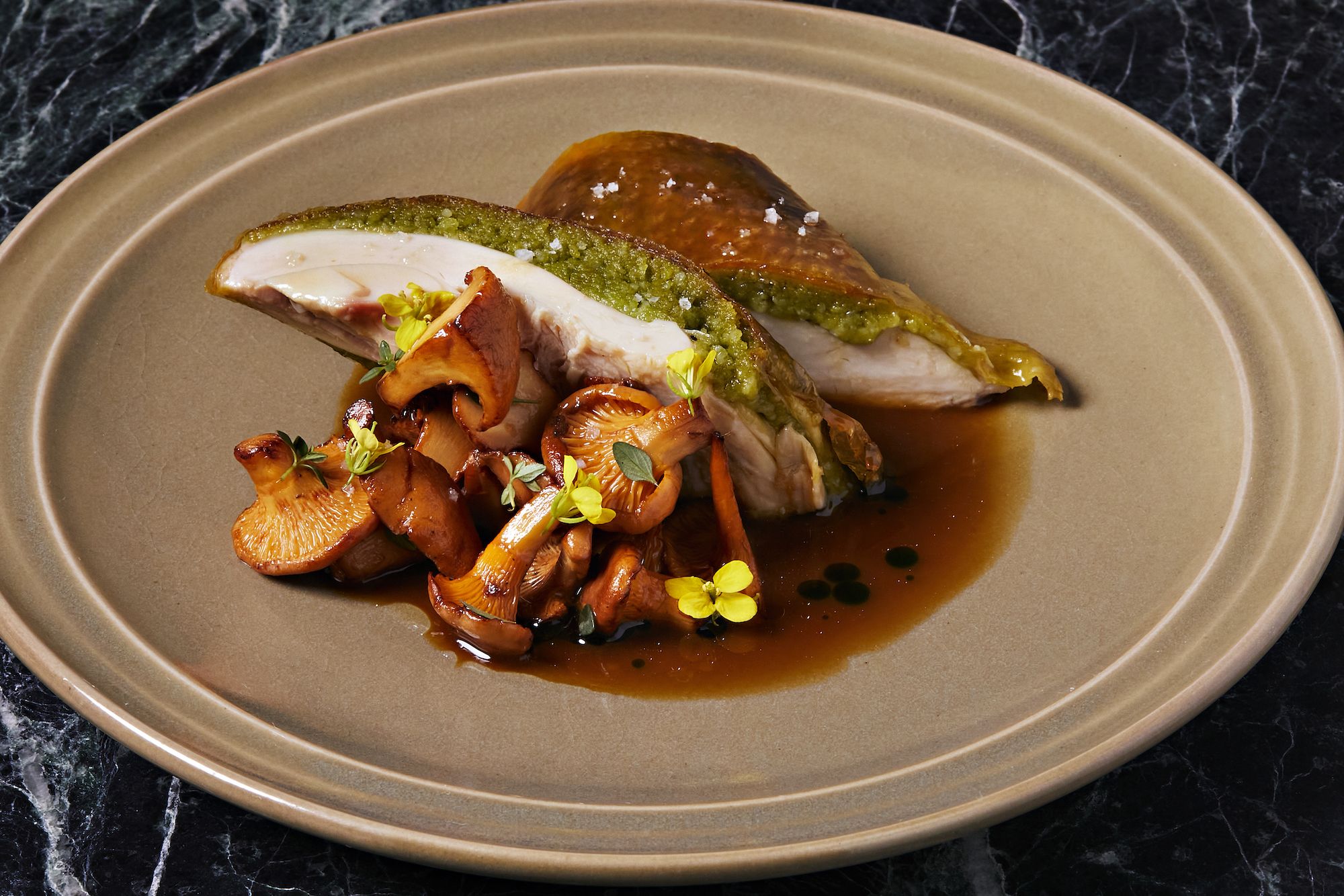 Two pieces of stuffed chicken with green herb filling on a tan plate, served with sautéed brown mushrooms, small yellow flowers, and brown sauce on a black marble surface.