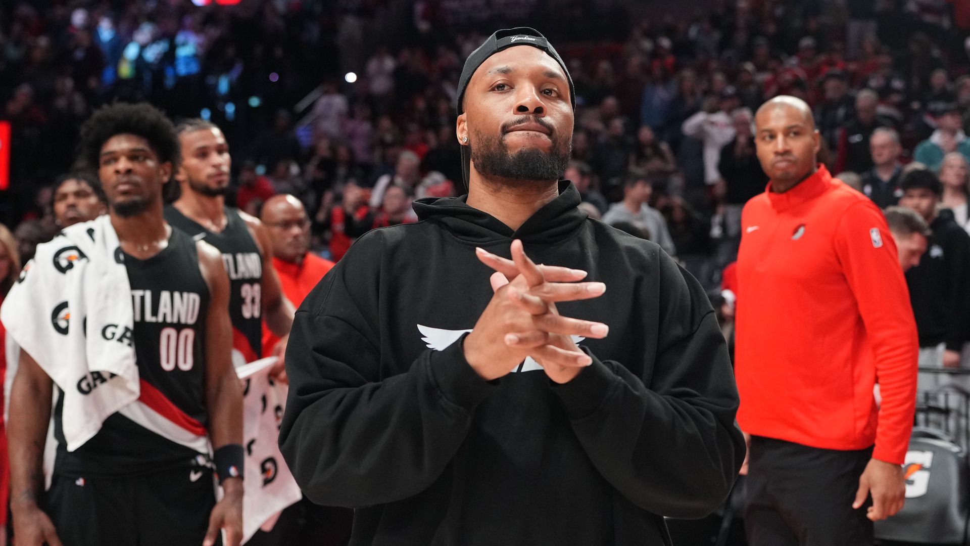 Close-up of a man in a black hoodie and backward cap on a basketball court, hands together. Behind him, players in Atlanta uniforms and a crowd in a packed arena.