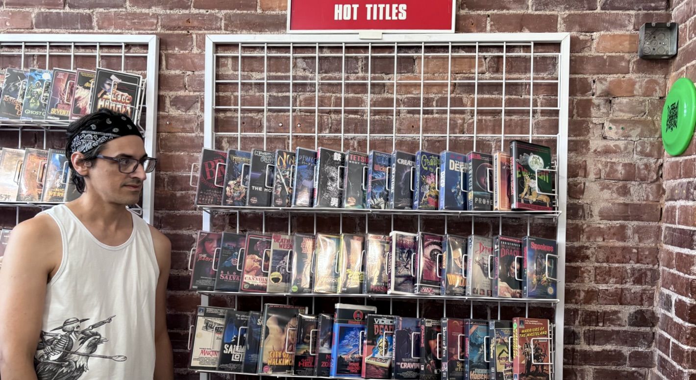 A man in a white sleeveless shirt and black bandana stands beside a brick wall, looking to the left, beside metal racks filled with colorful comic book covers; a red sign above reads HOT TITLES.
