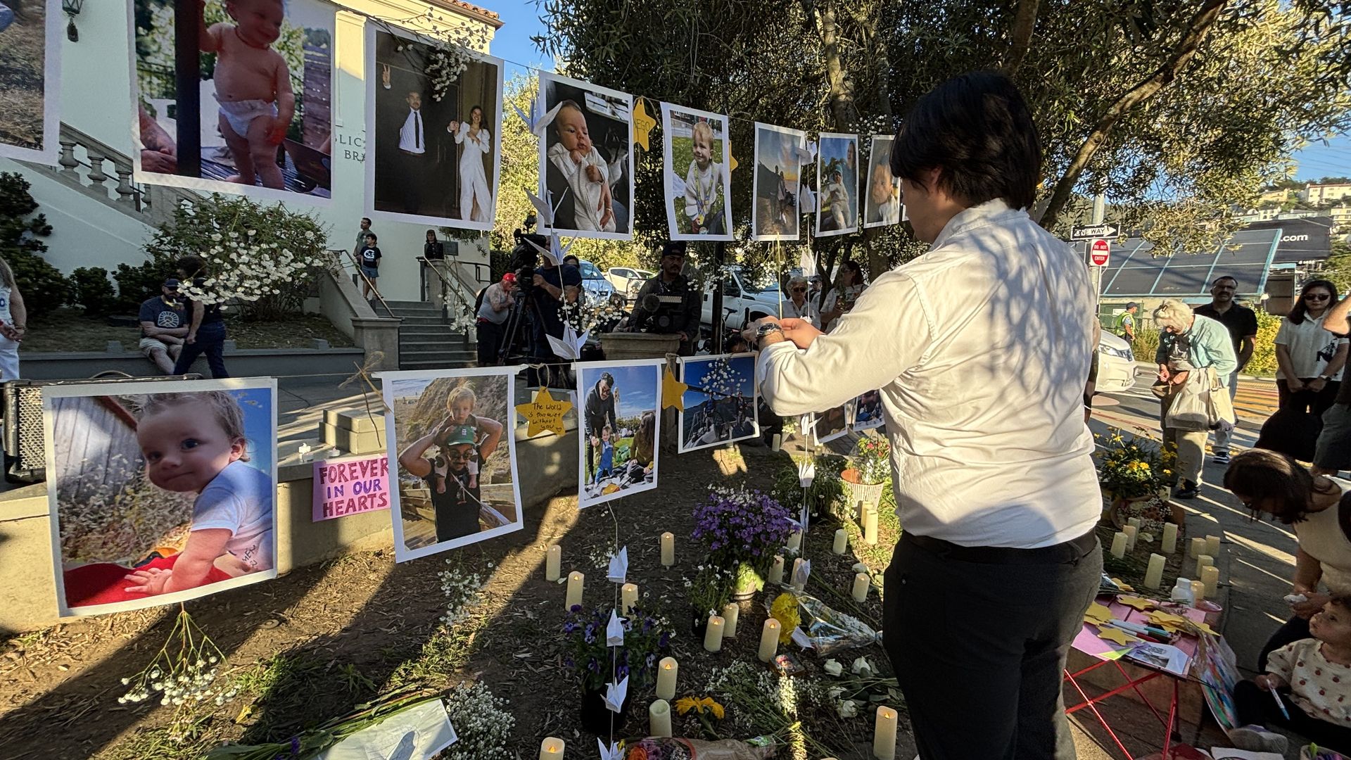 Outdoor memorial with large photos of babies and children hung on strings. Candles, flowers, and notes on the ground as people gather; a man in a white shirt adjusts the display.