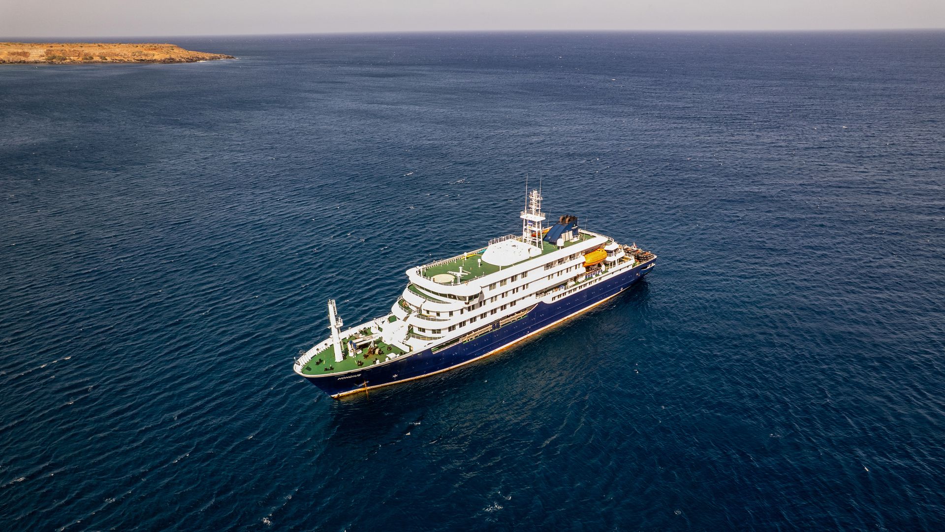 A cruise ship sits in dark blue ocean waters, with a light brown island nearby. 
