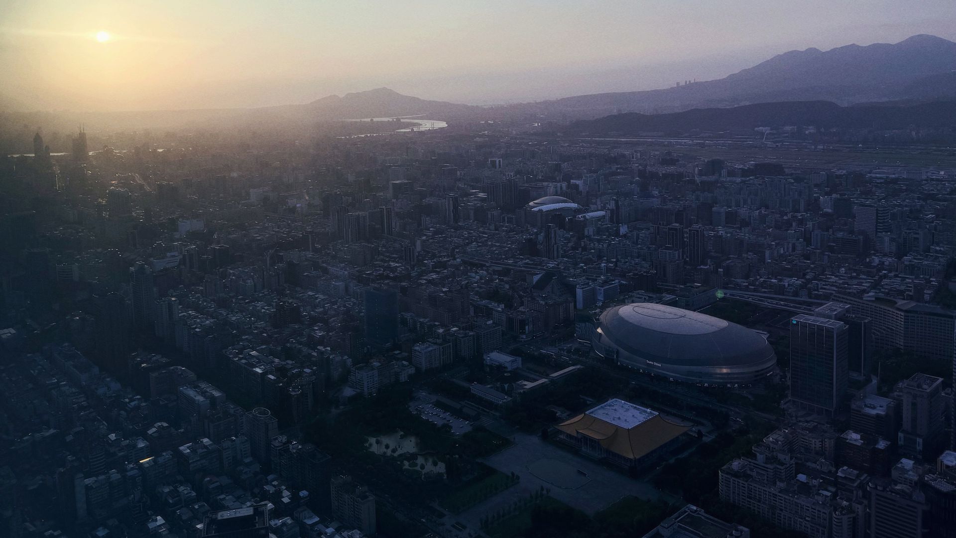 A cityscape at sunset with a hazy sky. The picture captures Taipei, the capital of Taiwan, with mountains in the background.
