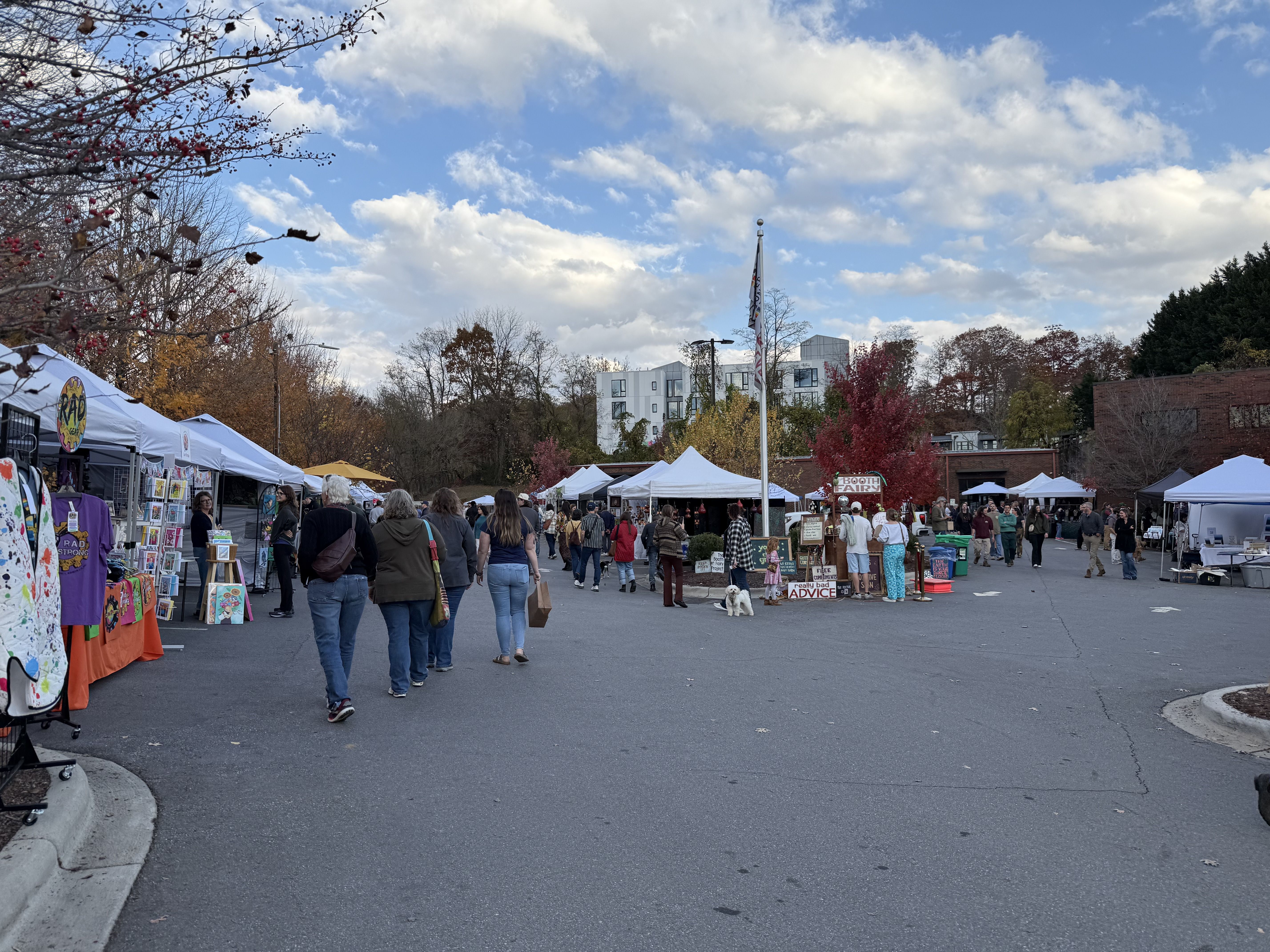 Outdoor market with people walking among white tents and colorful stalls on a cloudy day. Trees with autumn leaves and modern buildings visible in the background.