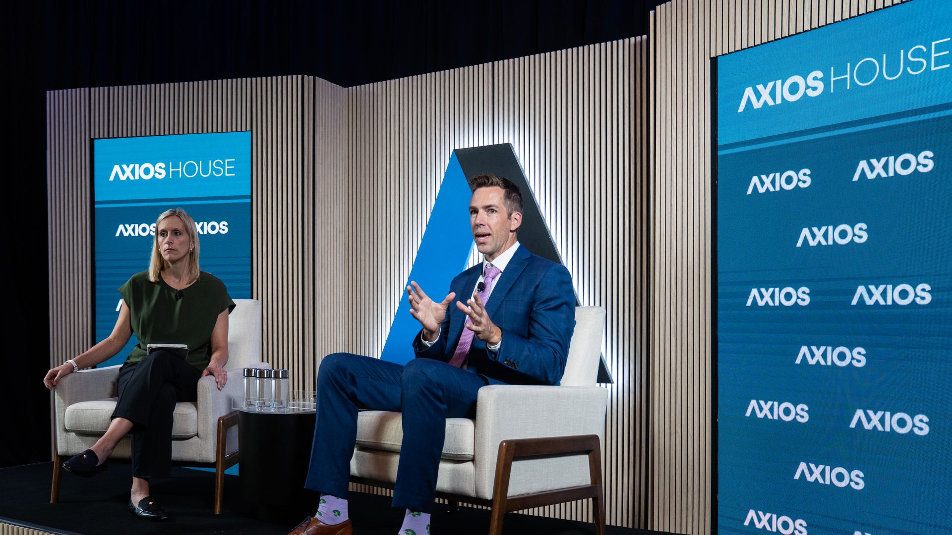 A man in a blue suit gestures to the audience while speaking. A woman sits in the background.