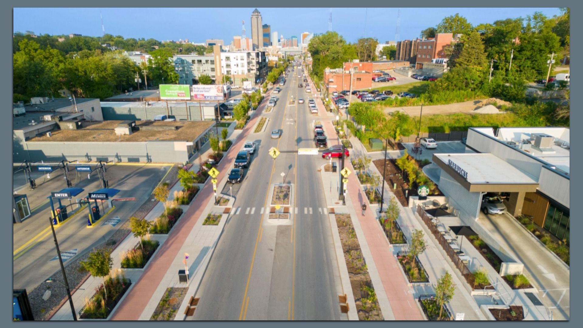 Aerial view of Ingersoll Avenue in Des Moines with cars parked on both sides, pedestrian crosswalks, sidewalks with trees, and a drive-thru Starbucks on the right under a clear blue sky.