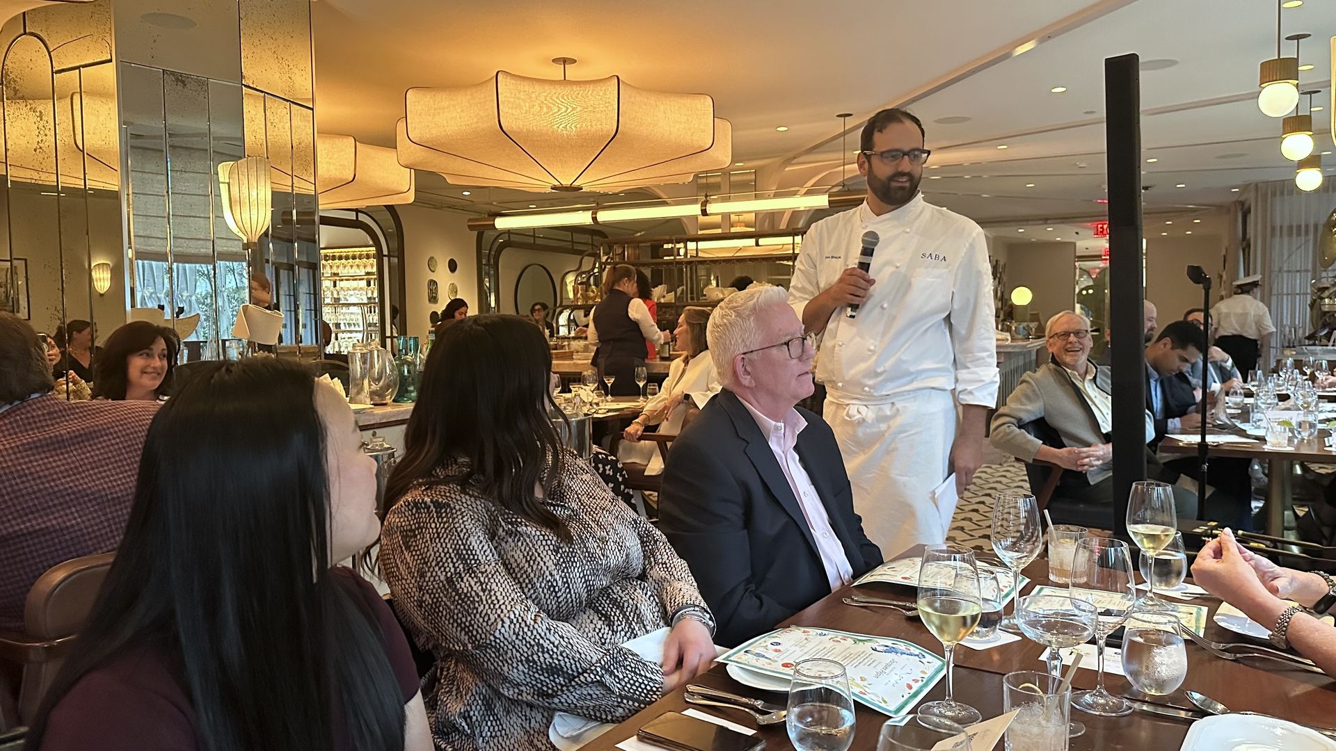 Chef Alon Shaya stands at center with a microphone as restaurant guests look to him.