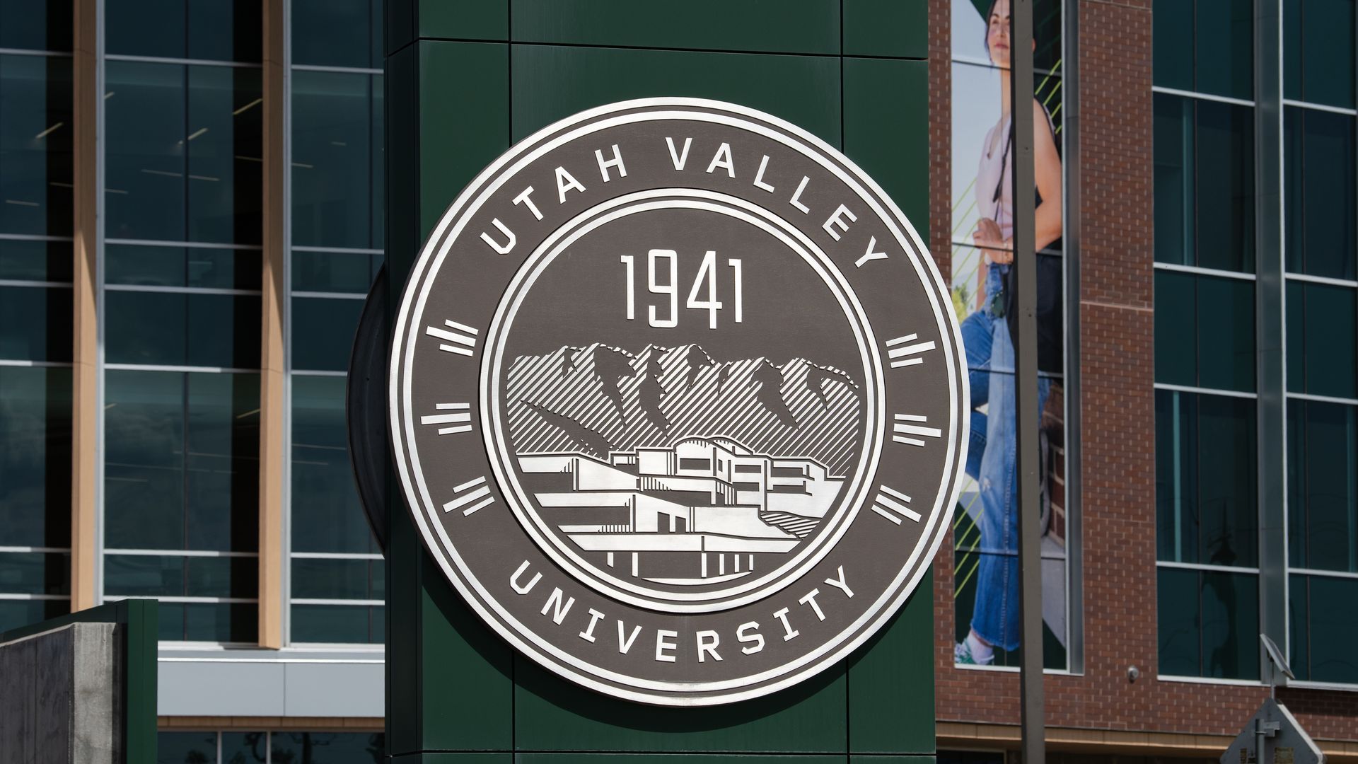 Close-up of a large circular Utah Valley University sign on a dark green pillar, with a glass-brick building behind and the year 1941 plus a mountain illustration on the sign.