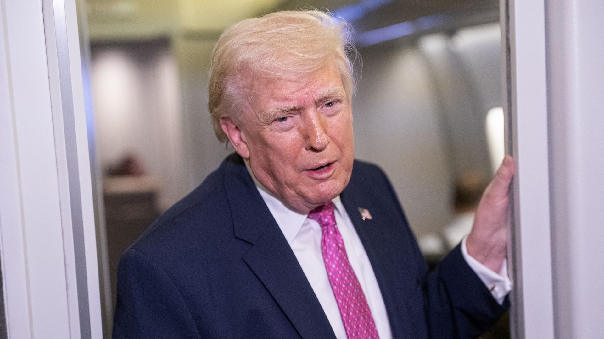 President Trump — wearing a dark suit, a white collared shirt, a pink tie and an American flag pin — leans through a doorway on Air Force One as he speaks to reporters, who are not pictured.