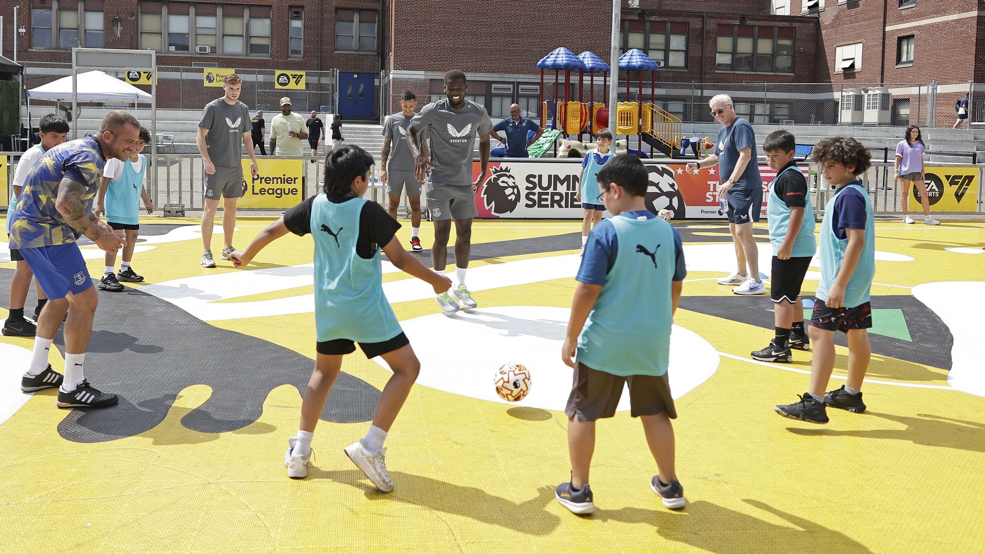Children playing soccer with adults wearing Everton gray shirts on a yellow outdoor court near a playground and brick school buildings under a blue sky.