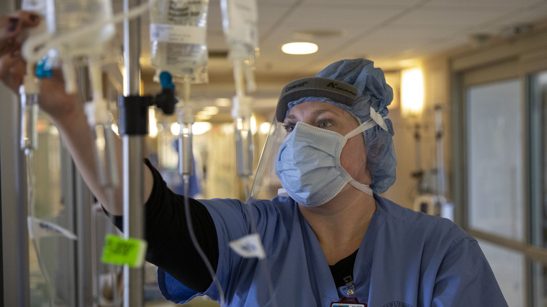 A nurse wearing a protective mask and shield monitors an IV.