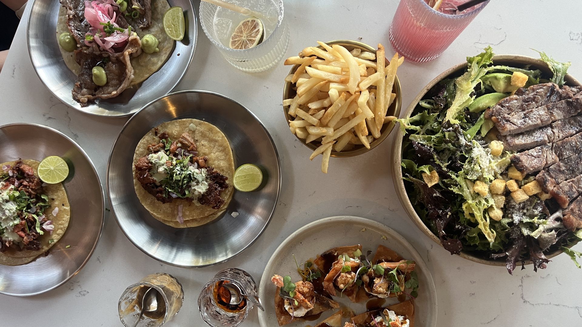 Top-down view of a shared feast: steak tacos with lime wedges, a bowl of fries, a large steak salad with greens and croutons, shrimp tostadas, a pink cocktail and a margarita.