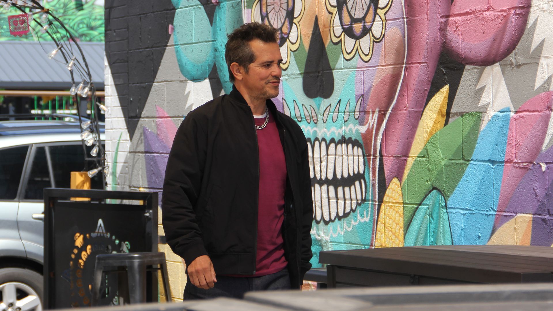 A man in a black jacket and a maroon shirt walks, while a colorful skull mural is painted behind him during a very sunny day. 