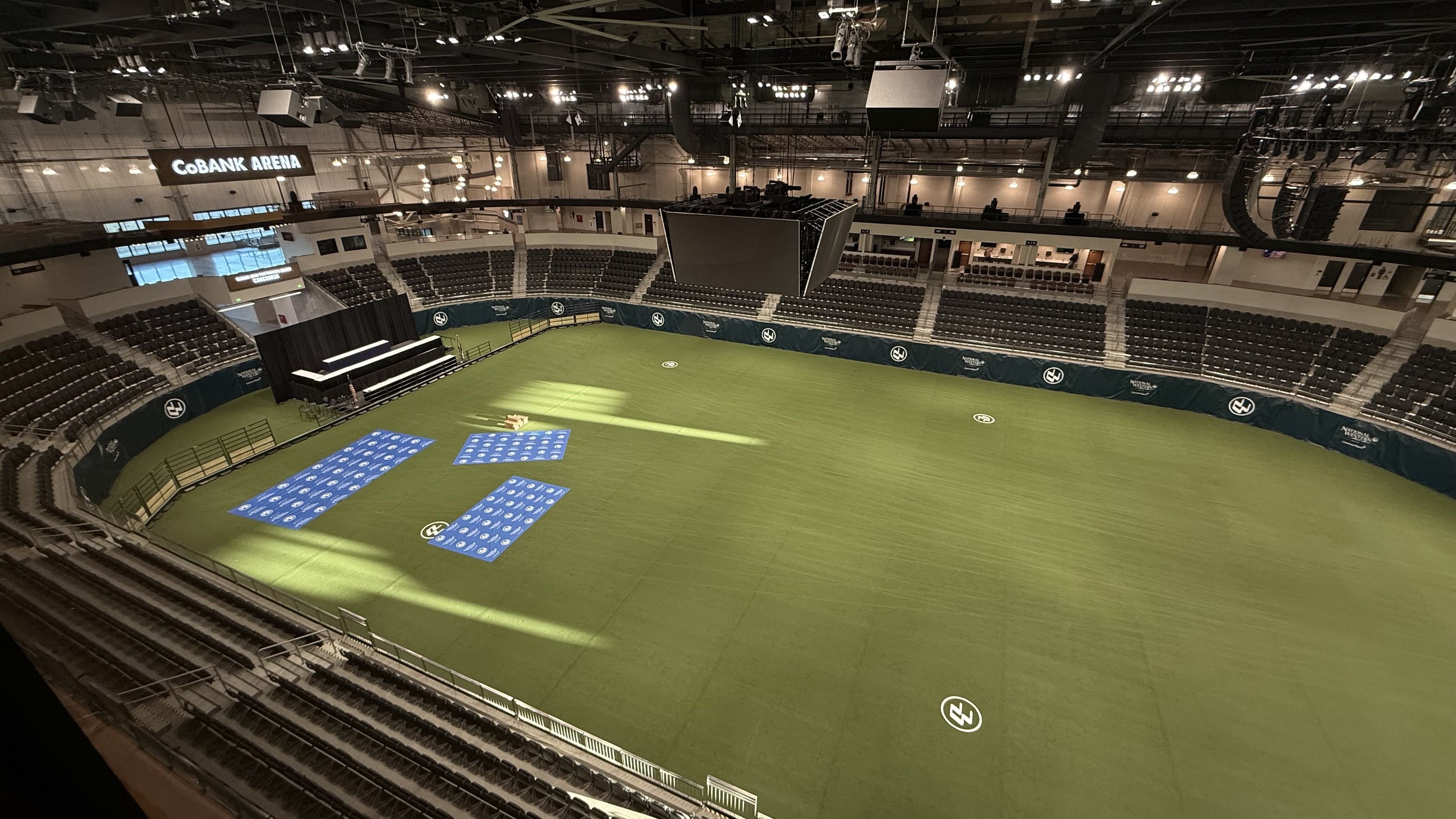 Interior of CoBank Arena with empty gray seats surrounding a large green floor marked with white logos and blue mats near a black stage setup with lighting overhead.