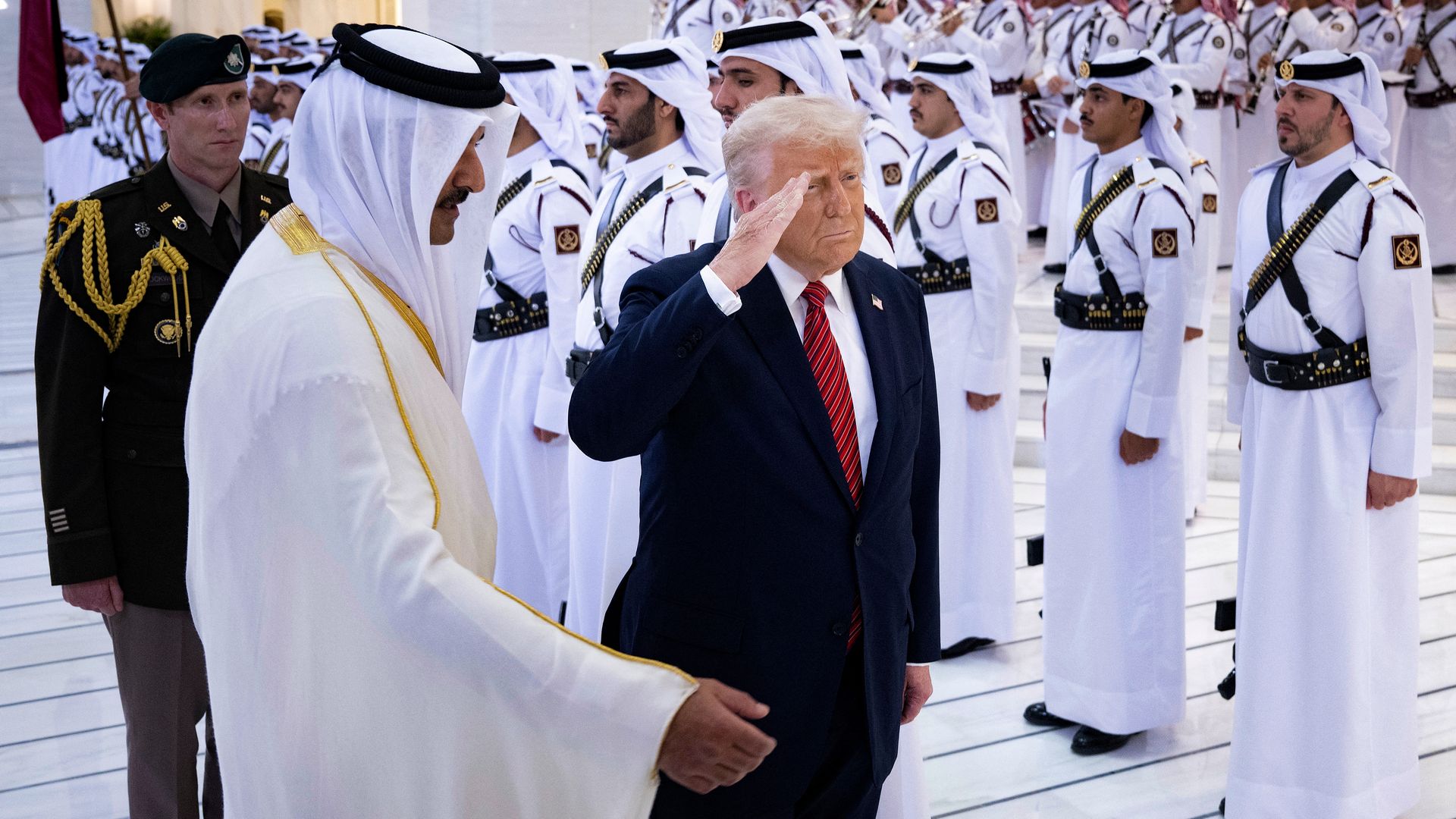 U.S. President Donald Trump, in a dark suit and red tie, salutes in a room full of men in Qatar. The frame is bright white.