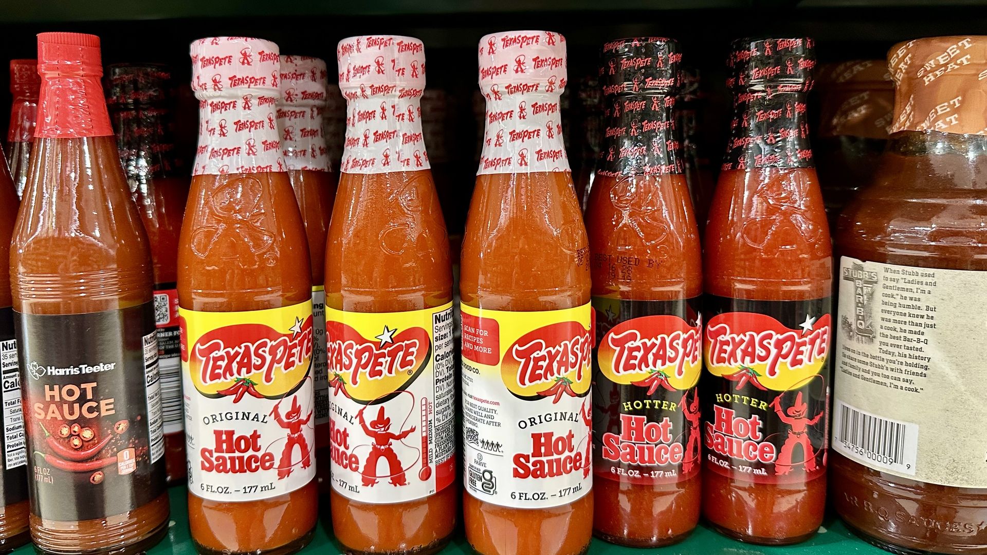 Row of Texas Pete hot sauce bottles on a green store shelf; clear glass bottles filled with orange sauce, red caps on the left, white caps with red Texas Pete logos in the center, bright display.