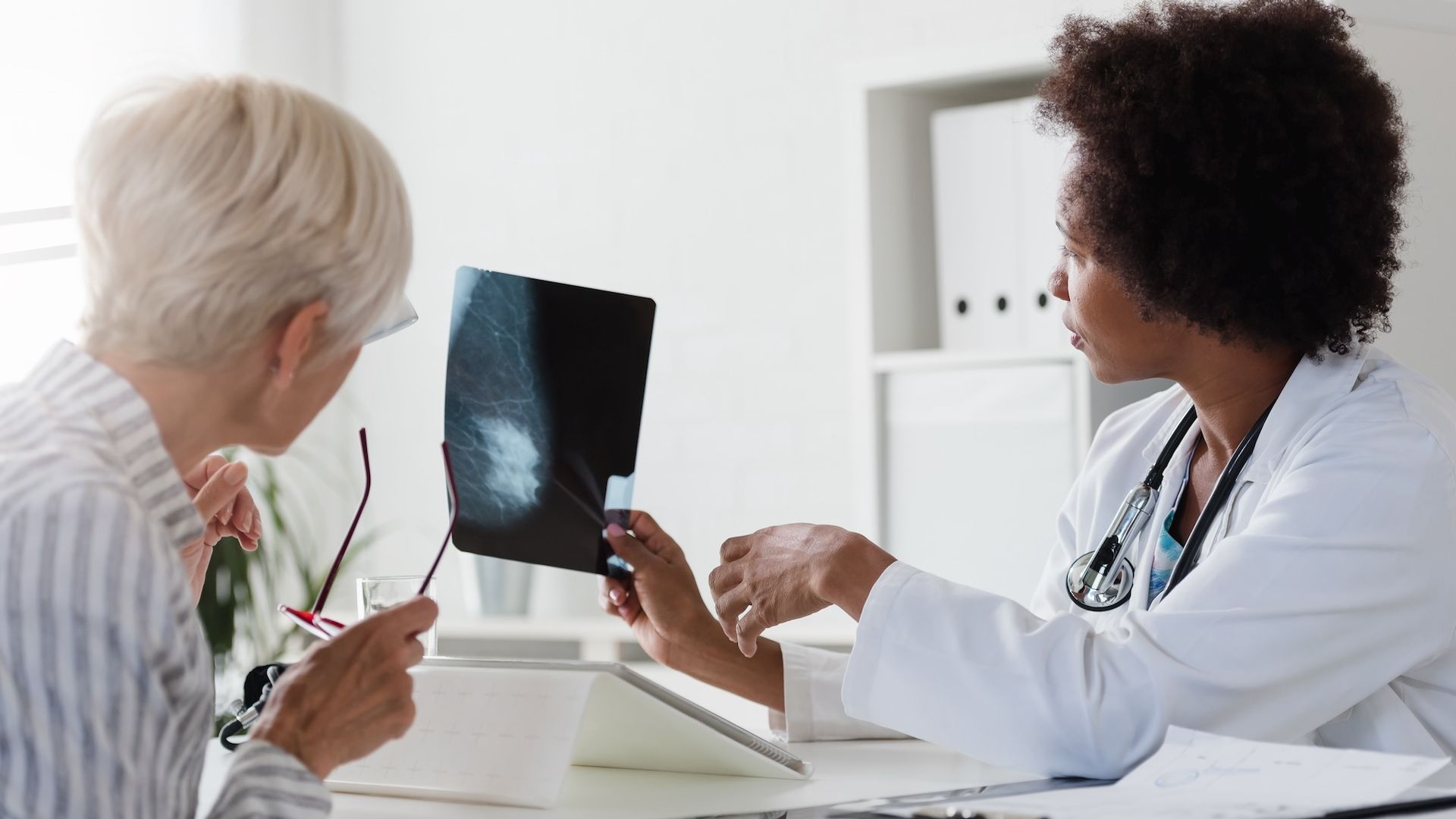 Female doctor with stethoscope showing a black and white mammogram image to an older woman with short white hair, who is holding her glasses attentively in a bright medical office.