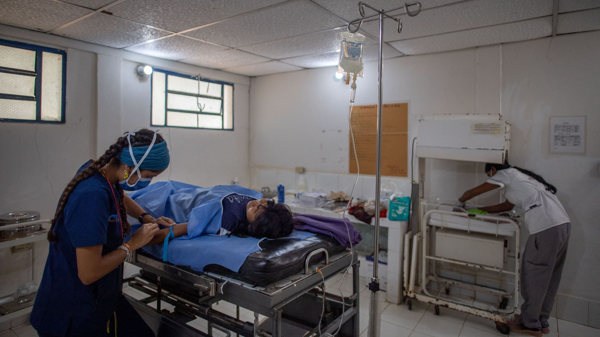 A medical staffer in scrubs tends to a woman laying on a hospital bed after she has delivered a baby. In the foreground, another person tends to a baby on a cart. 