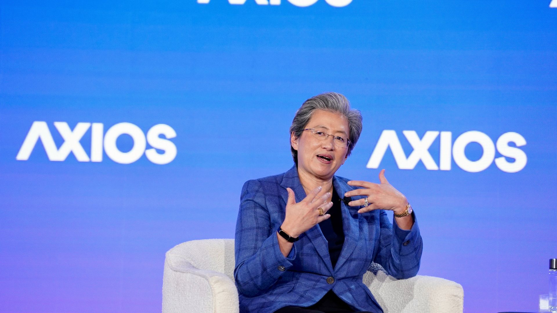 Person with gray hair wearing glasses and blue plaid blazer speaking while seated on a white chair in front of a blue background with repeated "AXIOS" logos.