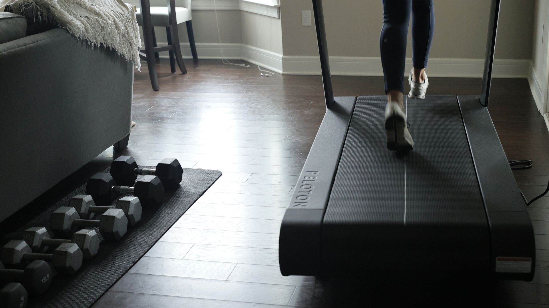 Photo of a person running on a Peloton treadmill indoors next to a mat with dumbbells 