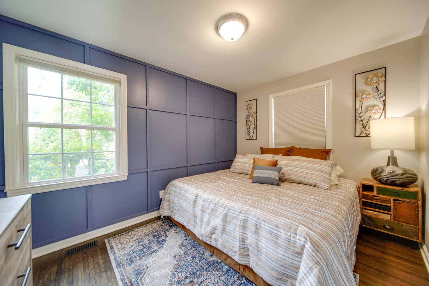 Cozy bedroom with a blue paneled accent wall, a bed with beige striped bedding and orange pillows, a wooden nightstand with a large lamp, framed floral art, window, and a patterned rug.