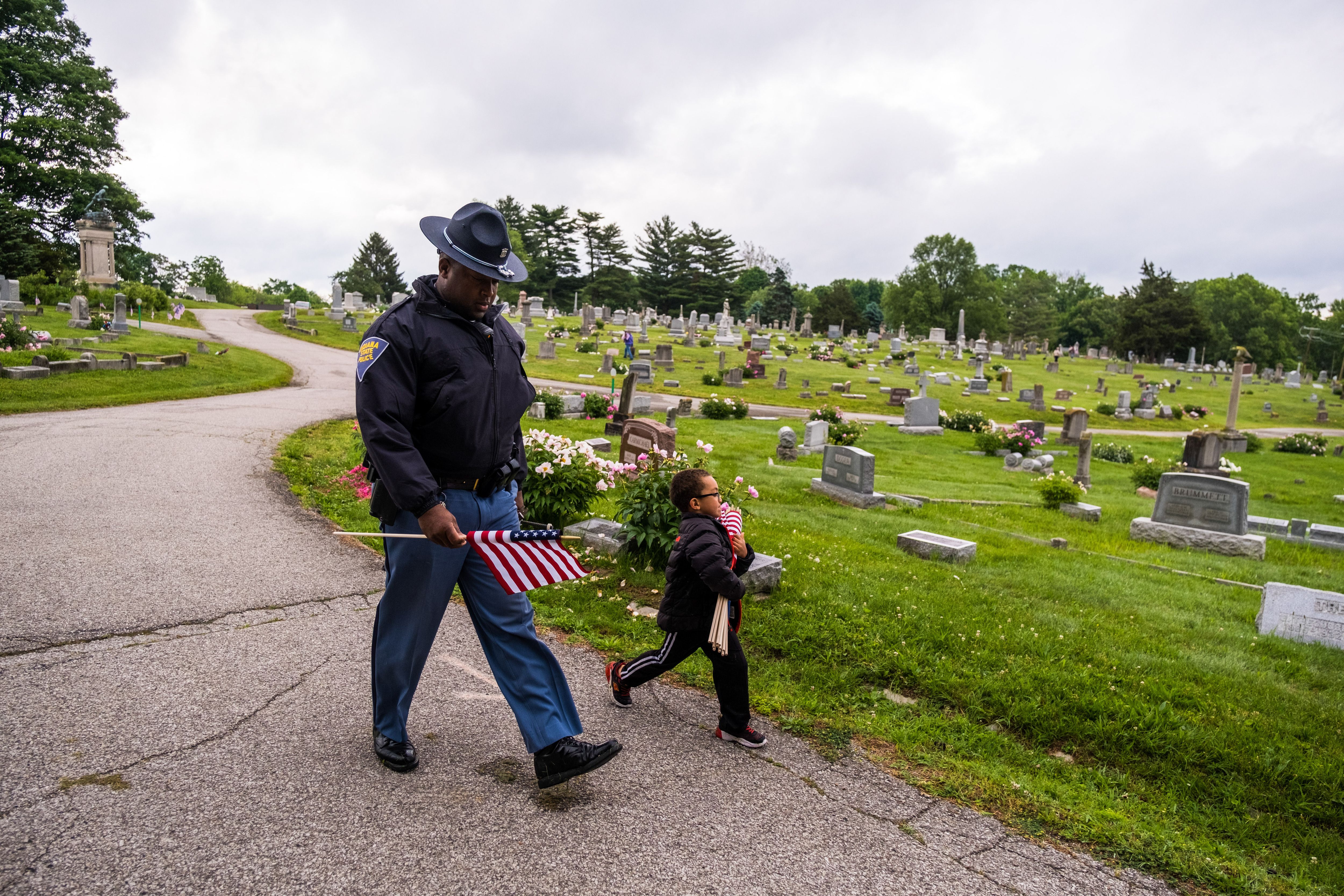 A state trooper walks down a path in a graveyard next to a child
