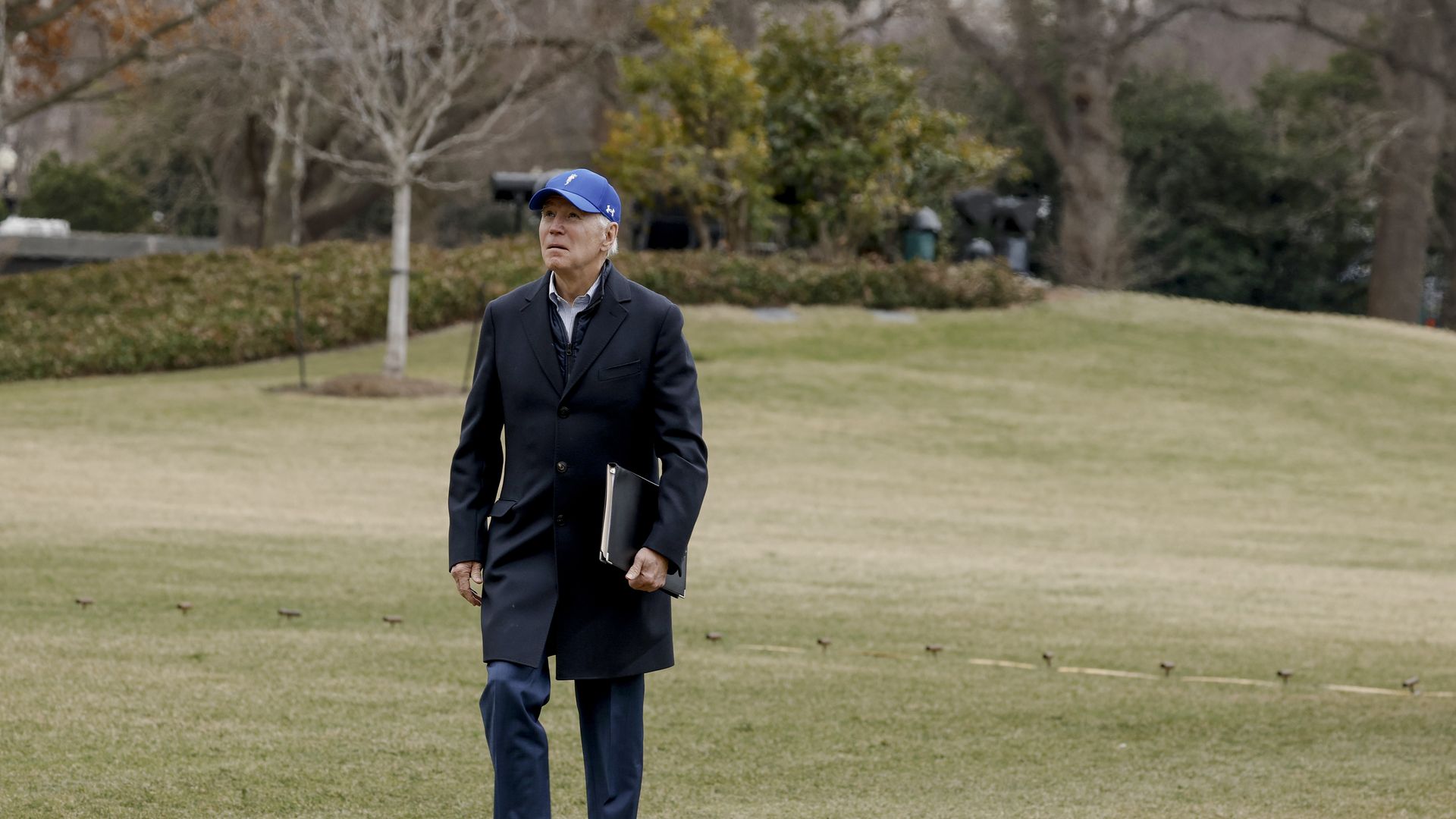 Joe Biden walks in winter grass on the White House south lawn, looking up the sky in his blue baseball cap