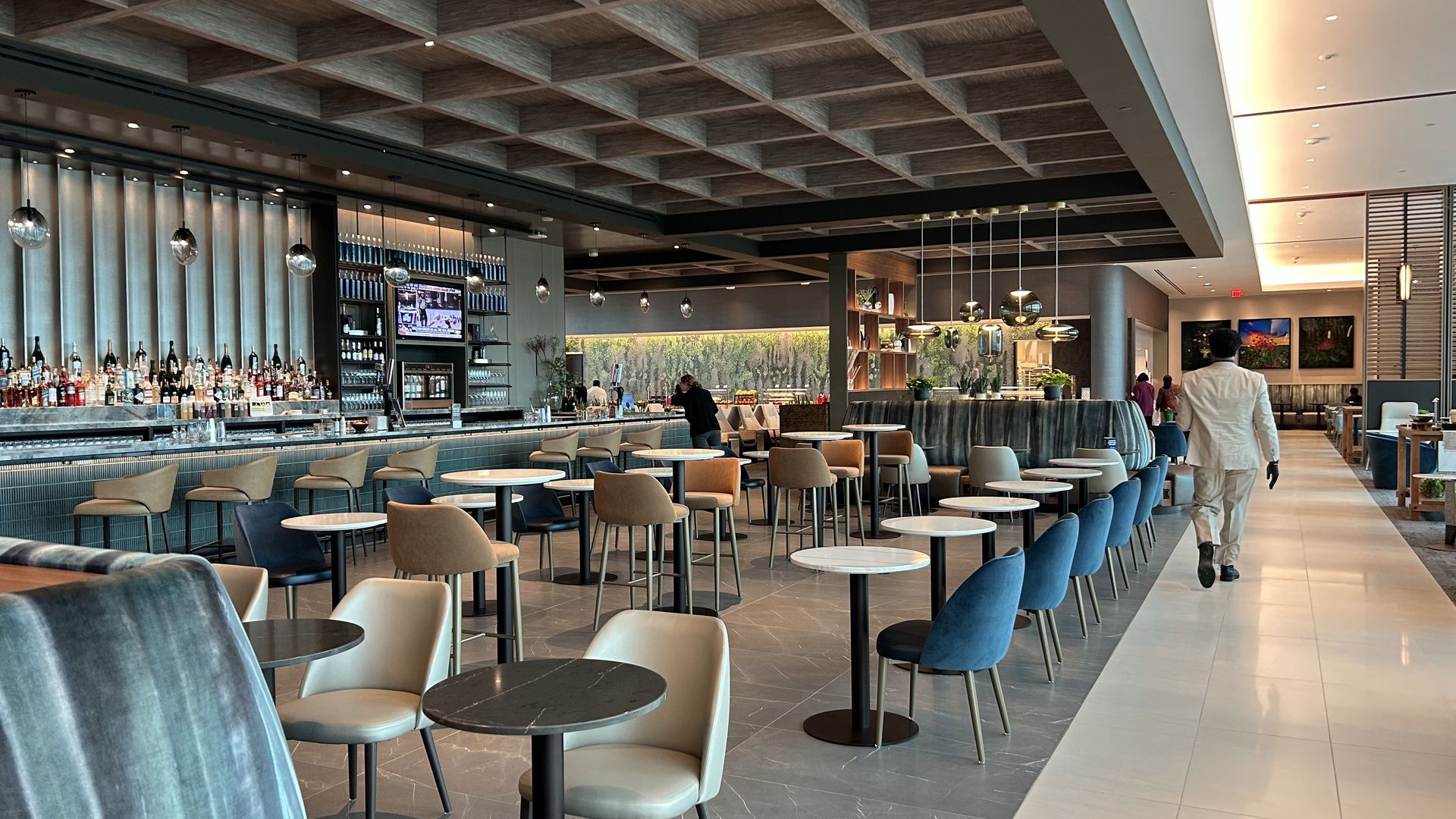 An individual in a suit walks in a large spacious exclusive airport lounge. A bar is along the left hand side of the lounge.