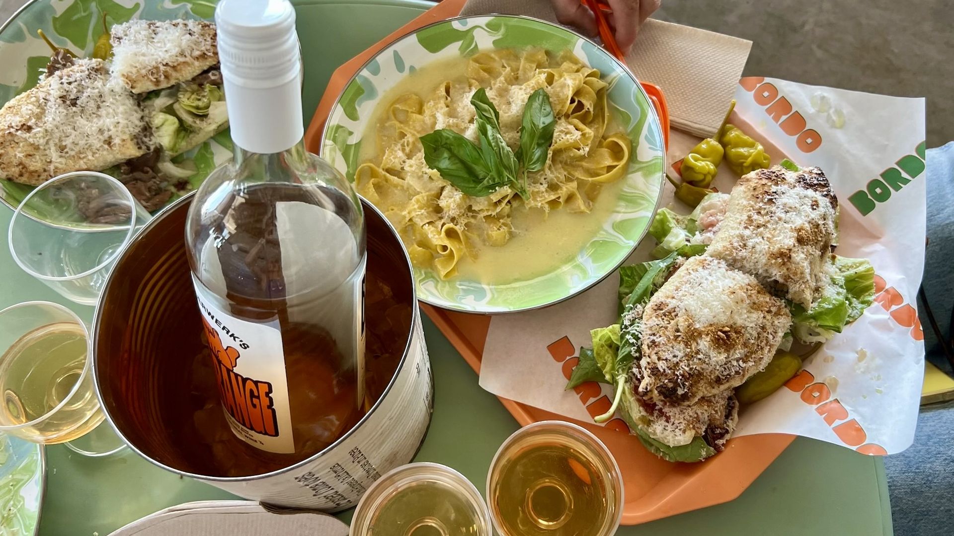 Top-down view of a bright green table with two plates: creamy pasta with basil, and breaded chicken croquettes on greens; a wine bottle in an ice bucket and several small glasses of white wine.
