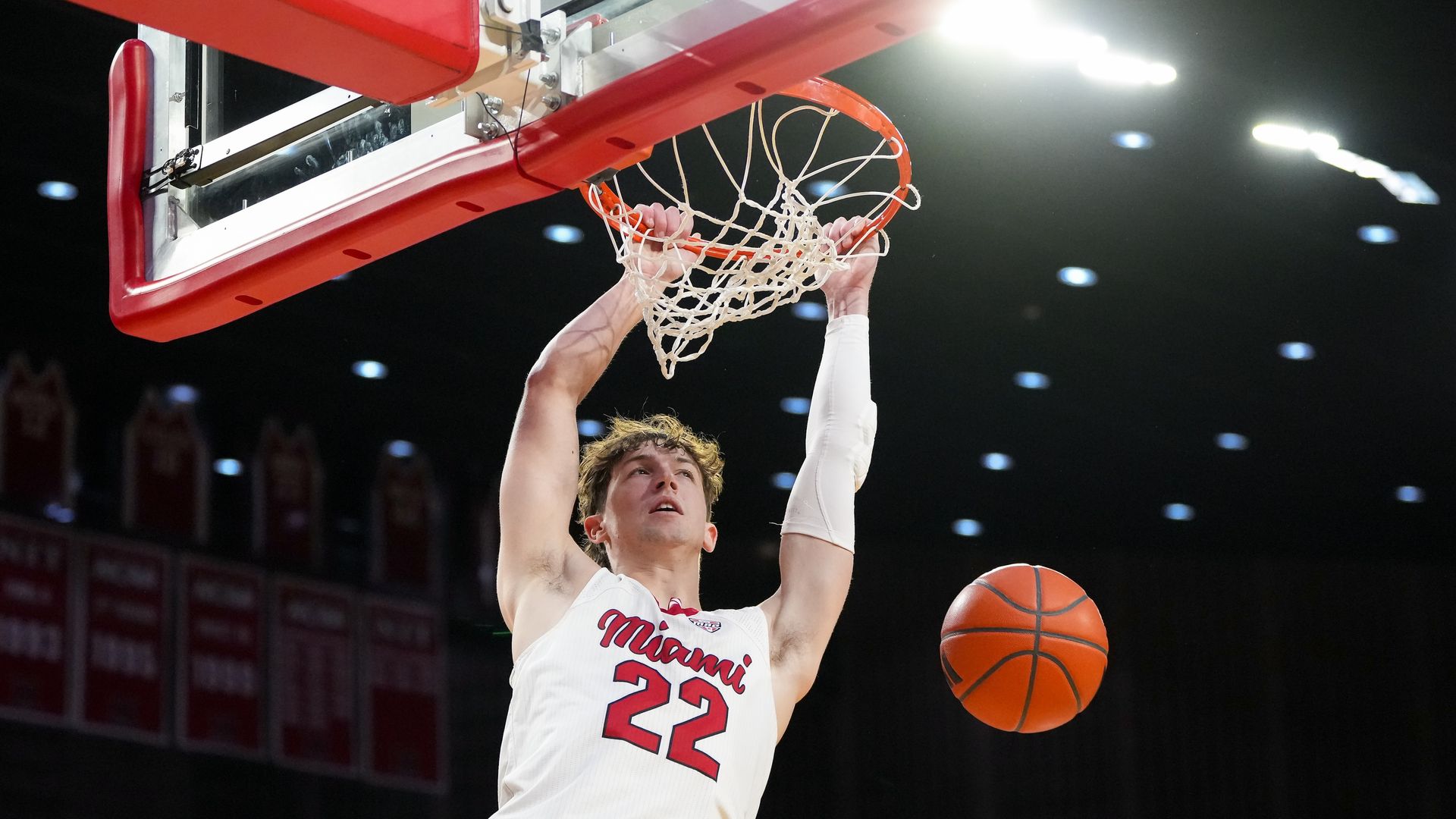 A basketball player in a white Miami jersey #22 dunks the ball through a red rim and white net; his left arm is taped. The arena lights and banners are visible in the background.