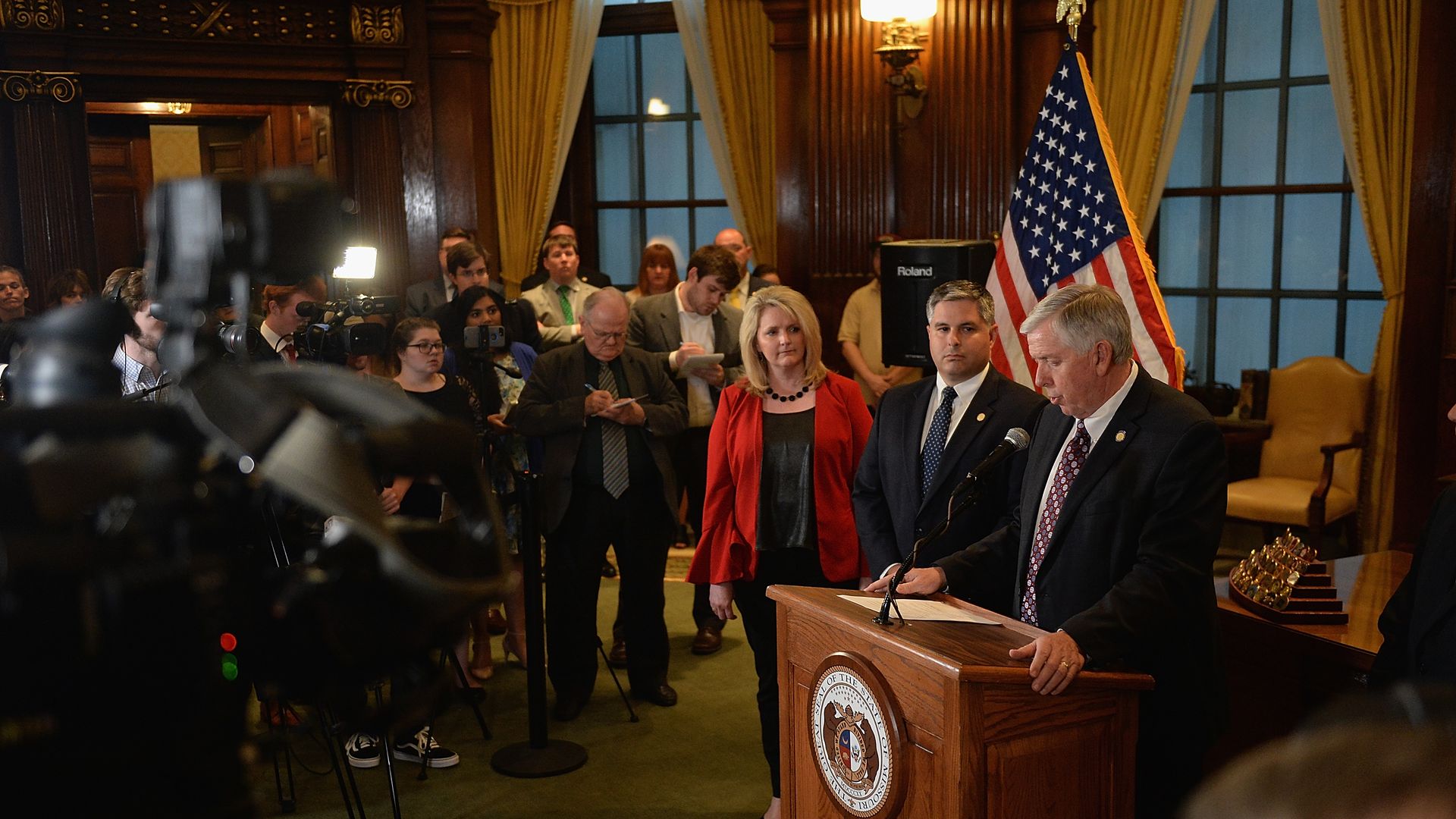 In this image, Michael Parson stands and speaks at a podium in front of a crowd and cameras.