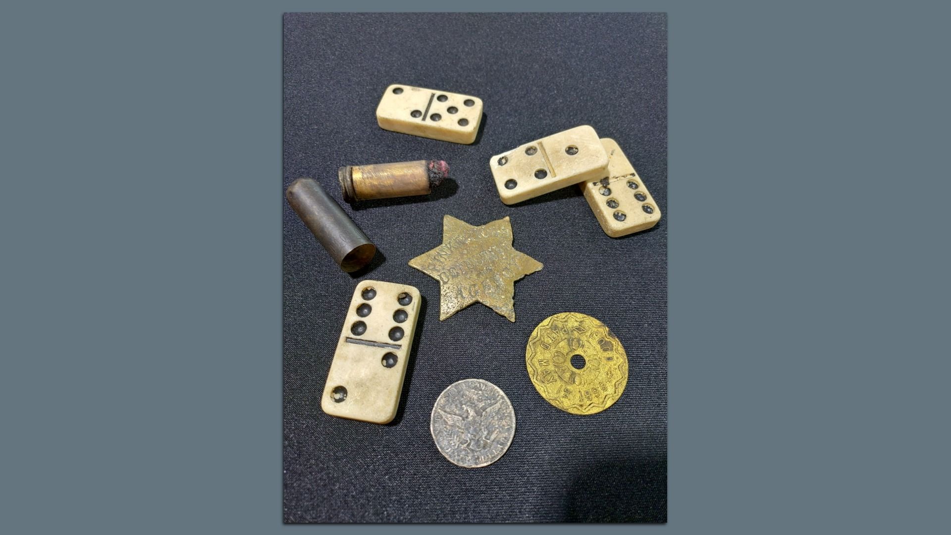 Vintage items on dark fabric: ivory domino tiles with black pips, a brass star-shaped badge, an old brass cartridge, a dark cylindrical tube, and silver and gold coins.