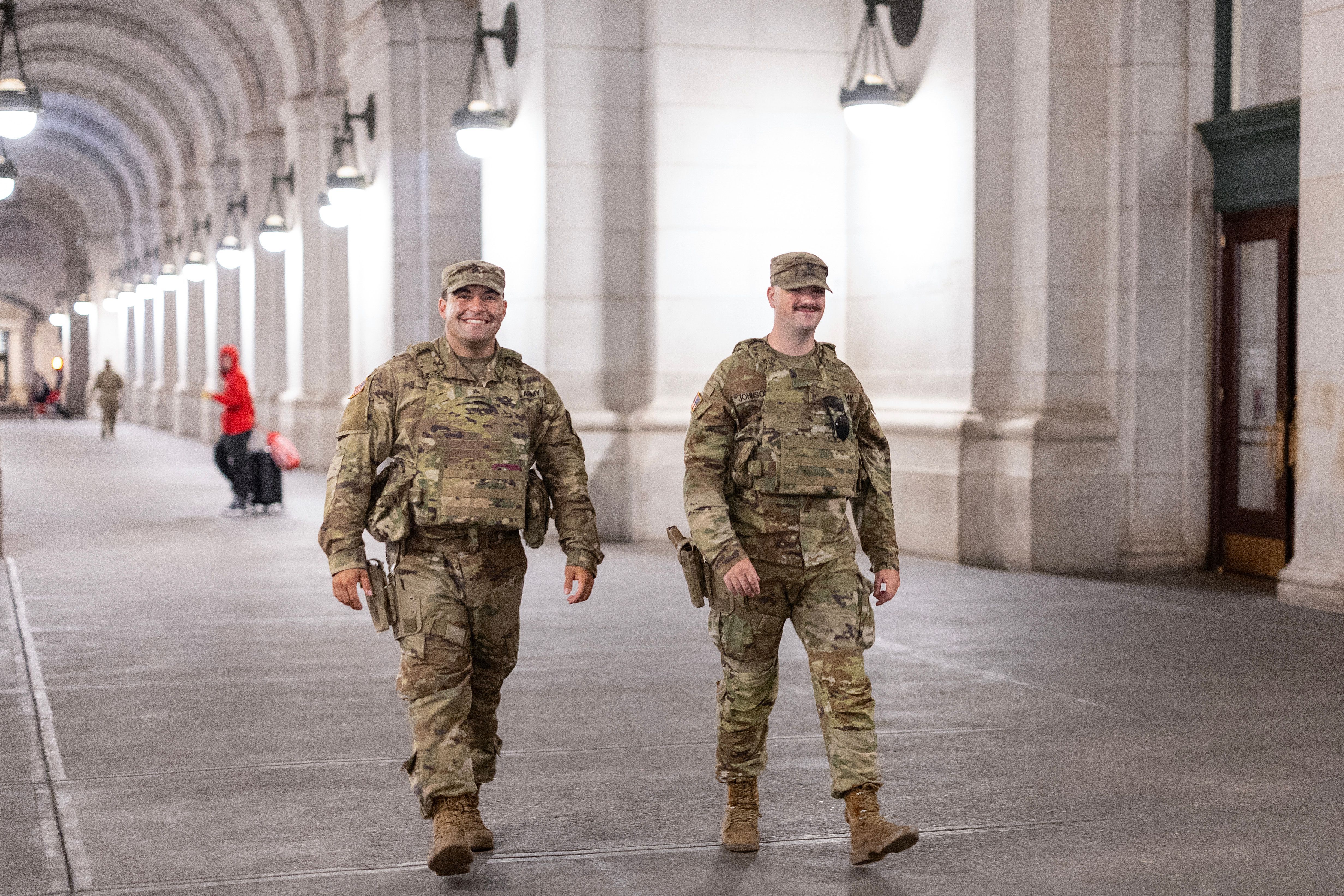 Two smiling soldiers in camouflage uniforms and tactical gear walking in a well-lit arched corridor with white walls and hanging lamps at night.