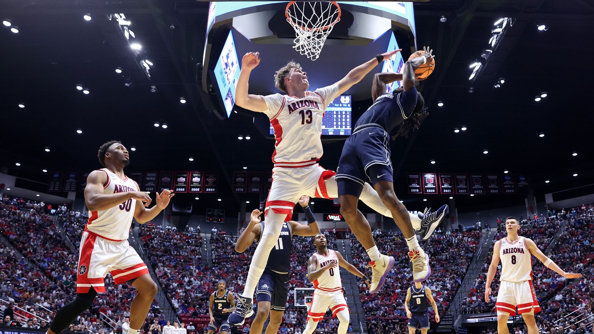 A basketball player in a white uniform with red striping on the side of his jersey  and the bottom of his shorts tries to block a layup attempt by a player in a dark blue uniform, while players from both teams look on in a packed arena. 