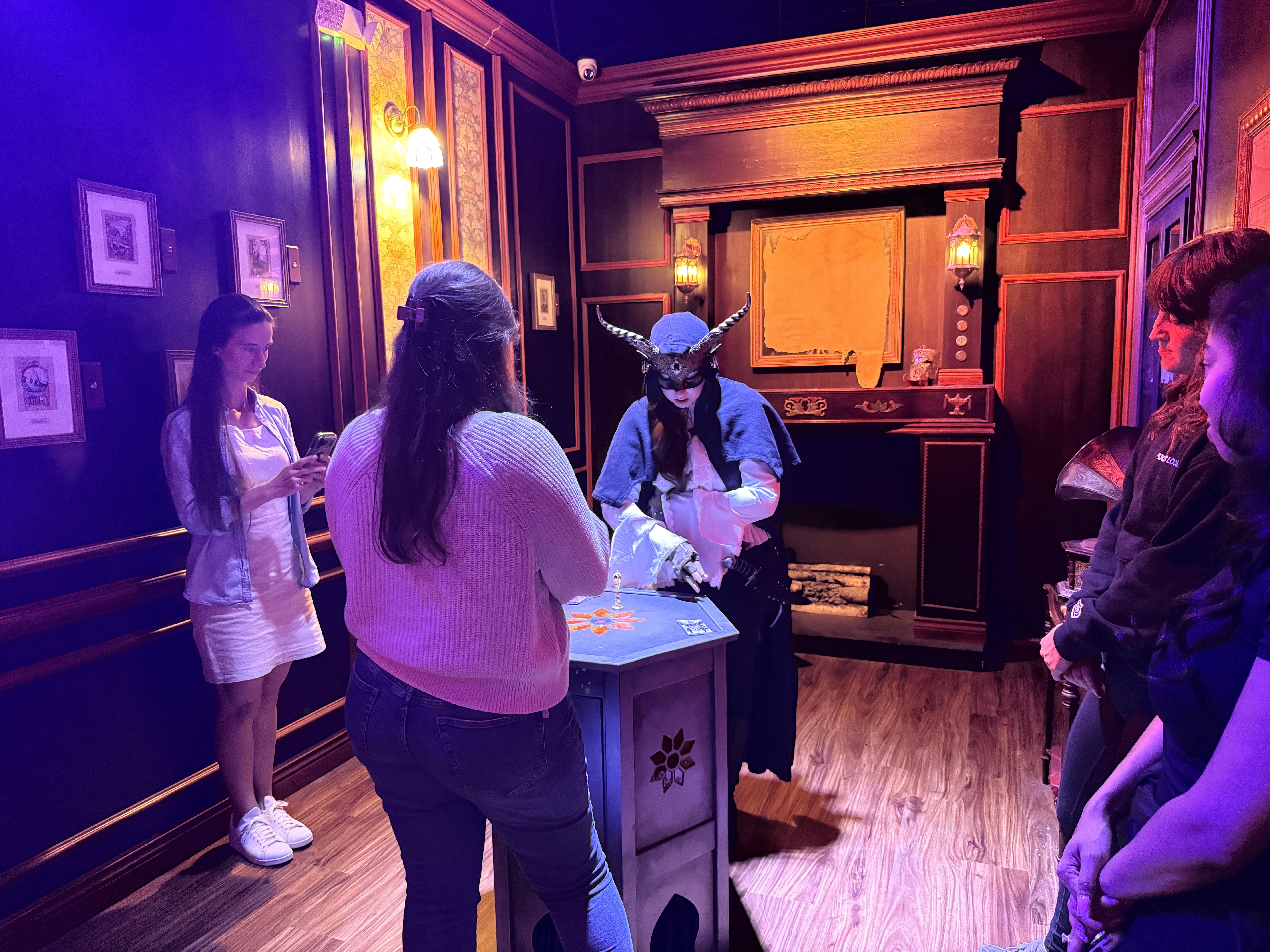 Dim purple-lit room with ornate wood paneling and framed pictures. A group of people gathers around a game table; a person wearing a horned mask and blue hood stands at the center while others watch.