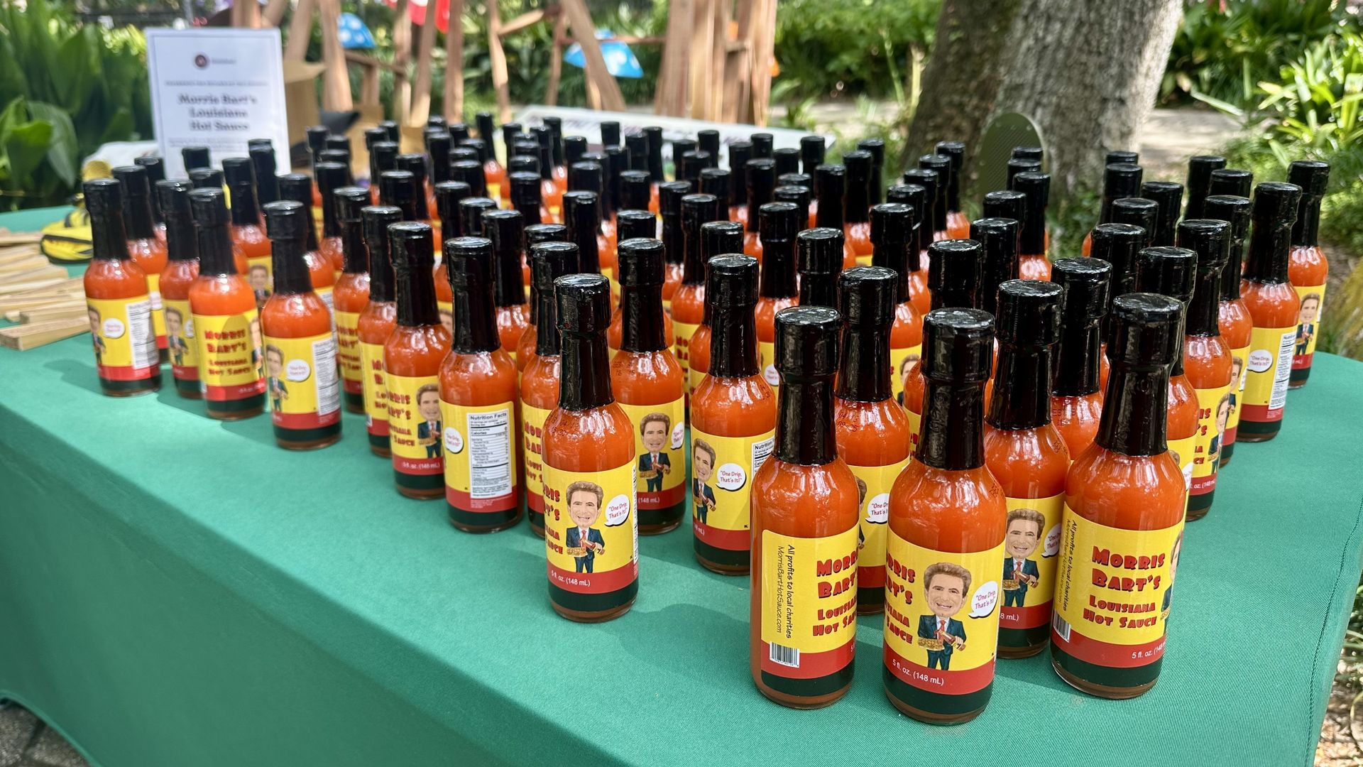 Outdoor display of dozens of hot sauce bottles with black caps and yellow labels on a green table; labeled Morris Bart's Louisiana Hot Sauce, arranged in rows with a blurred sign in the background.