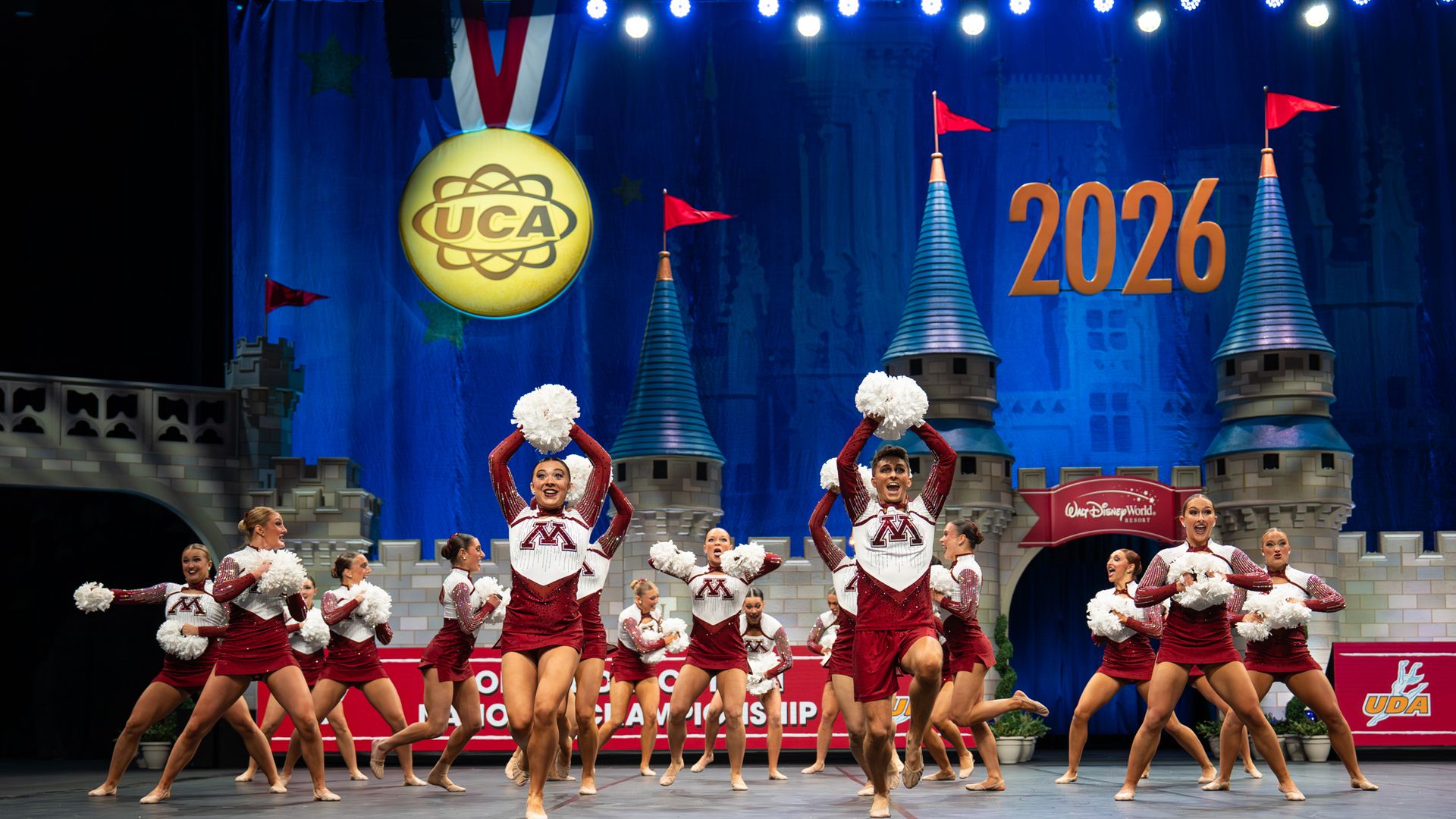 Cheerleaders in maroon and white uniforms with "M" perform with white pom-poms on stage decorated like a castle, with a large yellow UCA medal and "2026" in orange on blue backdrop.