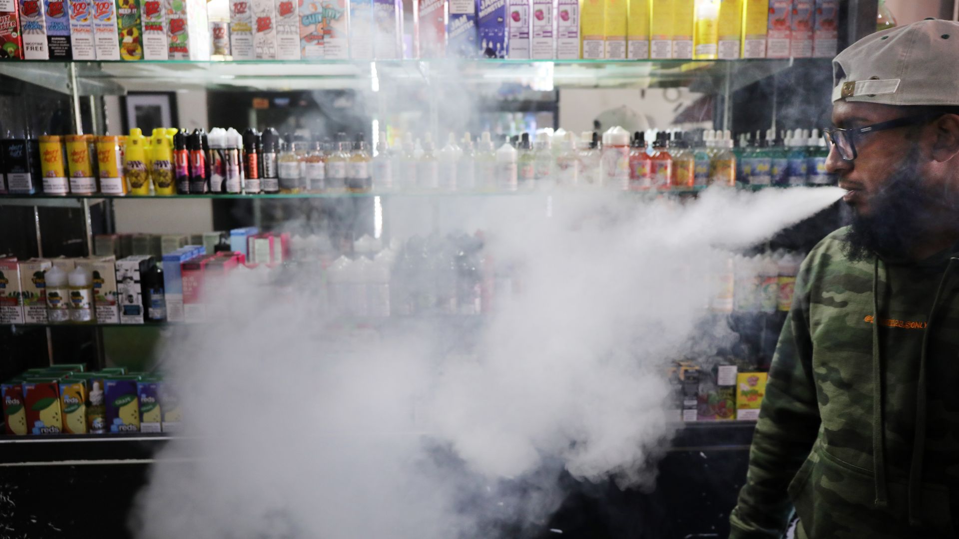In this image, a man blows smoke inside a vape shop, where the walls are lined with e-cigarette products.