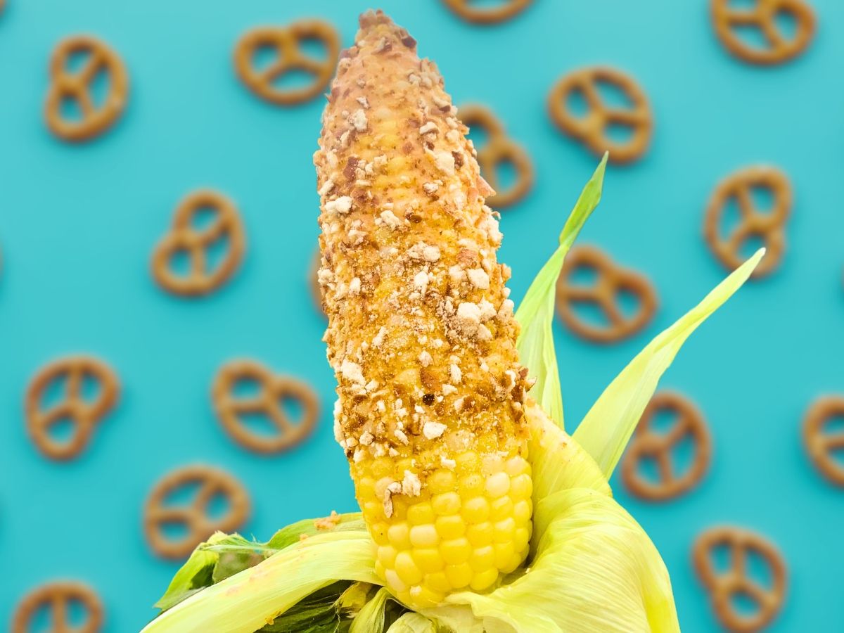 An ear of corn with brown-ish crunchy bits sticking to it in front of a blue backdrop patterned with pretzel illustrations.
