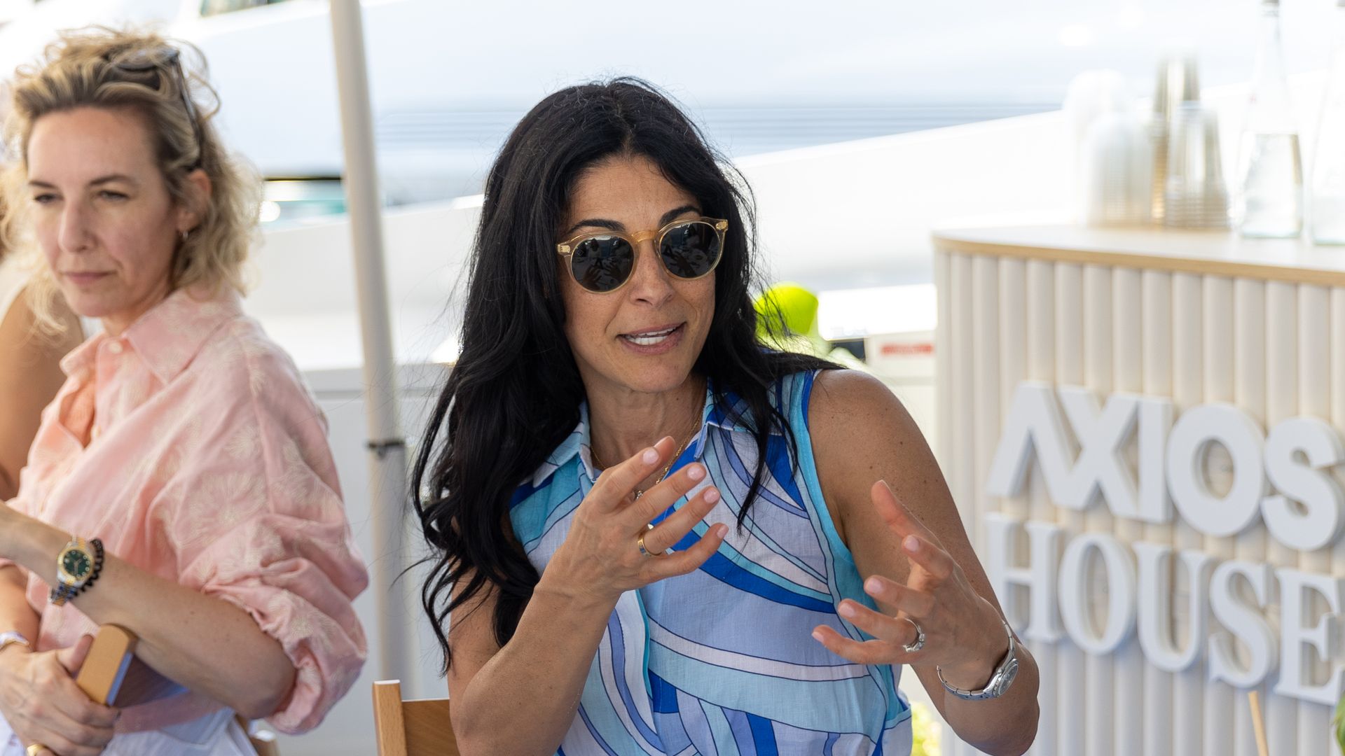 Two women sit at a sunny outdoor table. One in a blue striped sleeveless top with sunglasses gestures as she talks; the other in a pink blouse listens. Plates and drinks on the table.