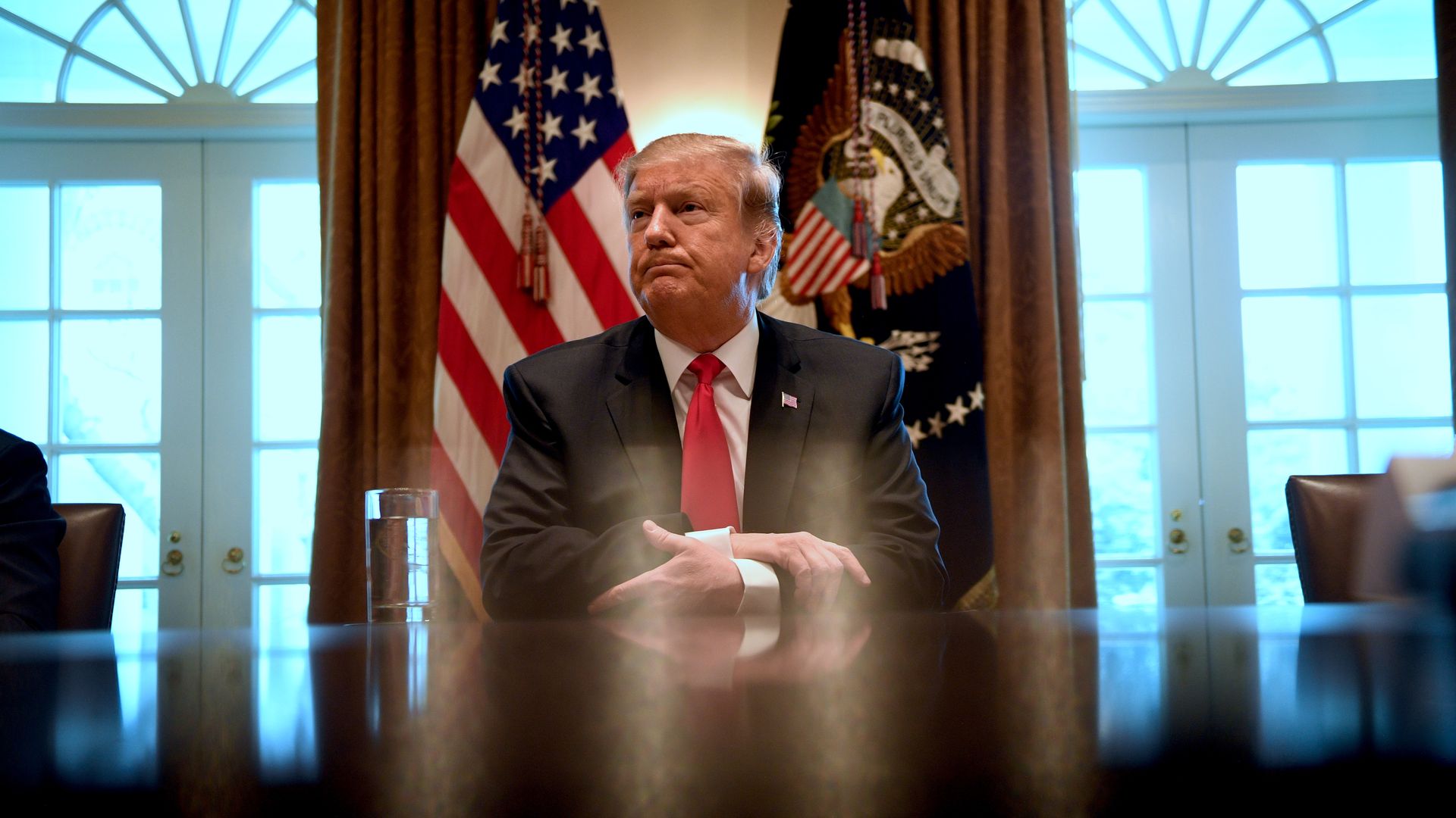 In this image, Trump sits at a desk with his hands folded and listens to someone out of view speak. There are two flags behind him.