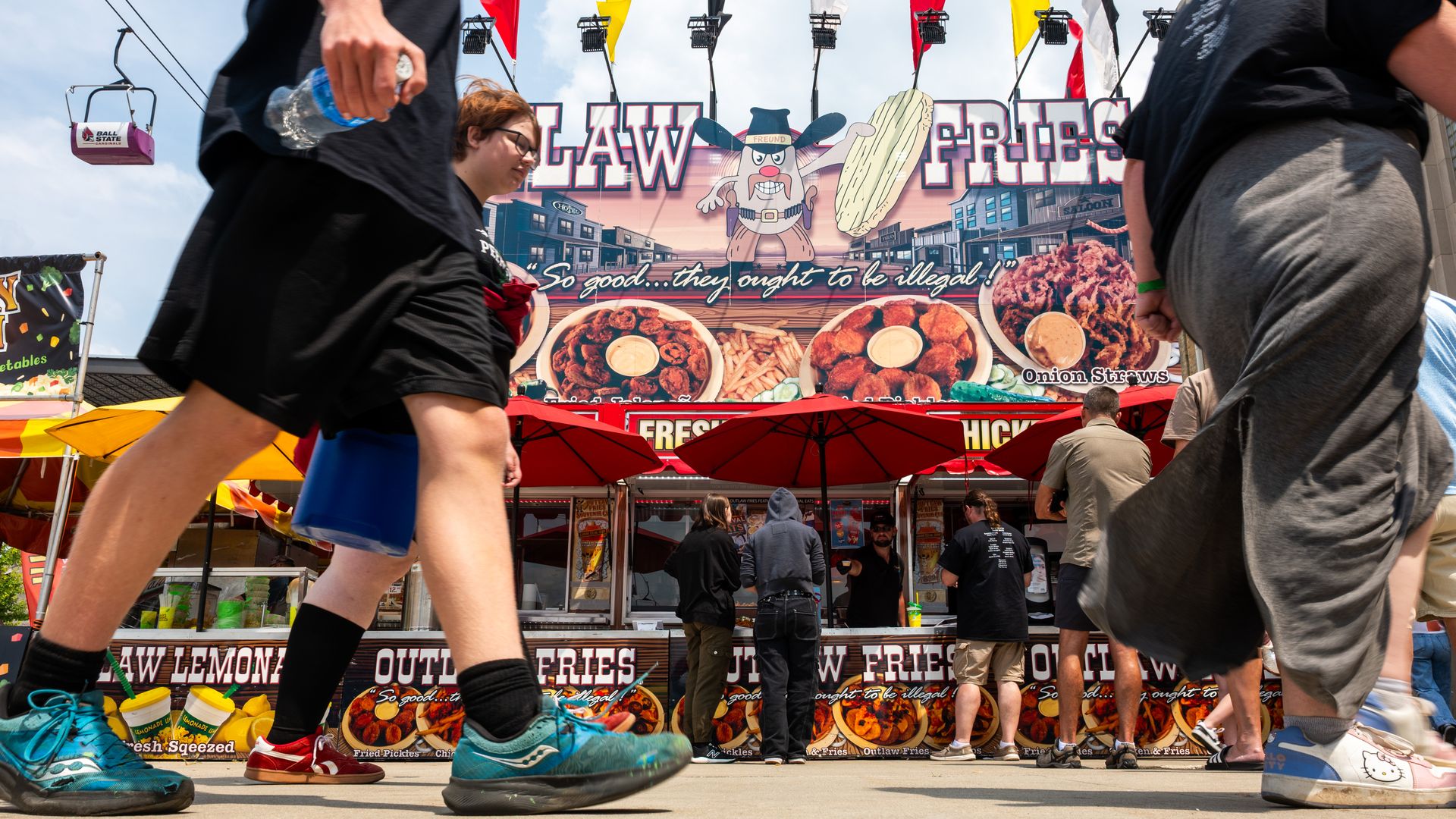 People walking and standing in line at a colorful food stand with a large sign advertising Outlaw Fries, featuring fried pickles, onion straws, and the slogan. 