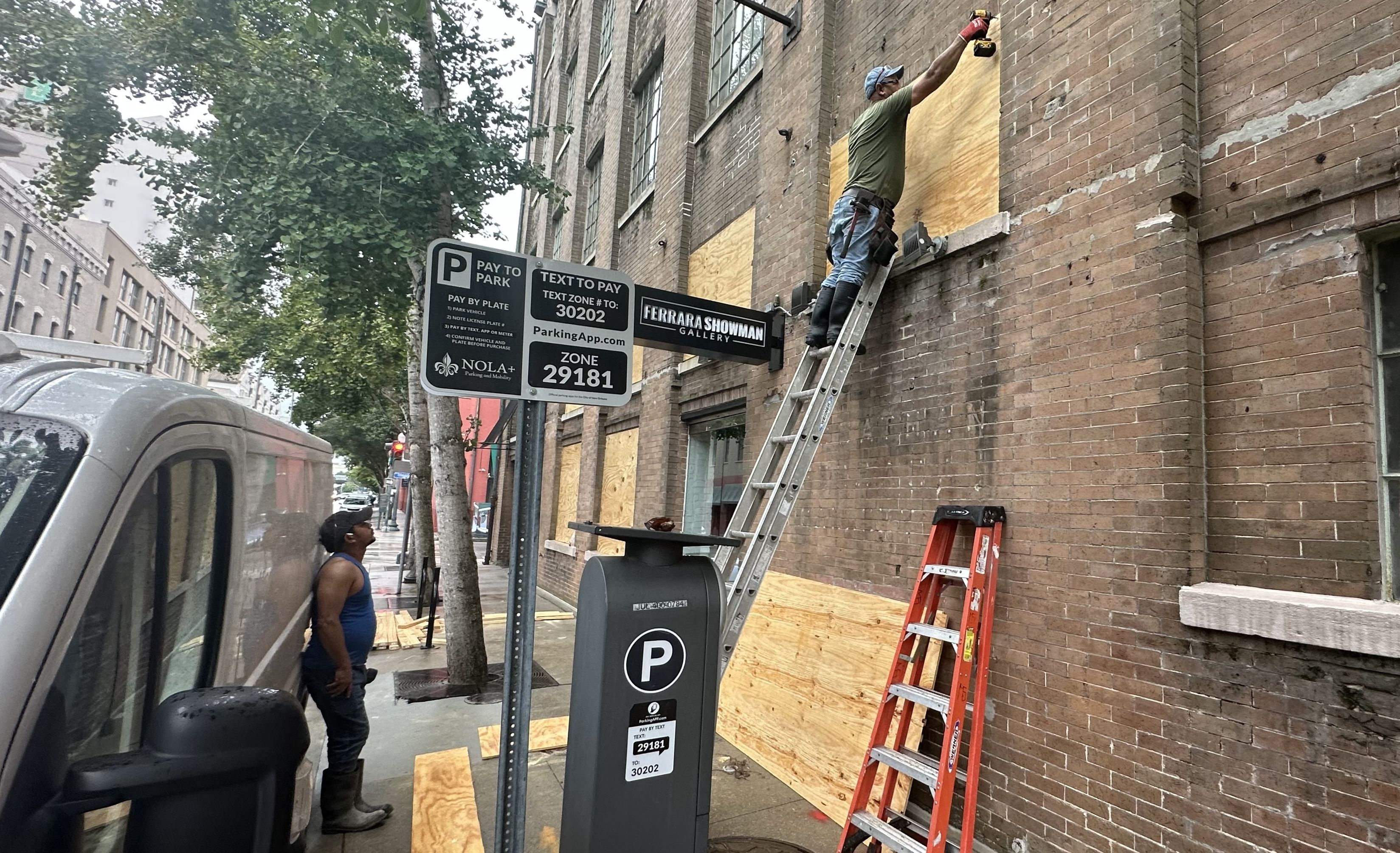 A man stands on a ladder as he screws a board over a window.
