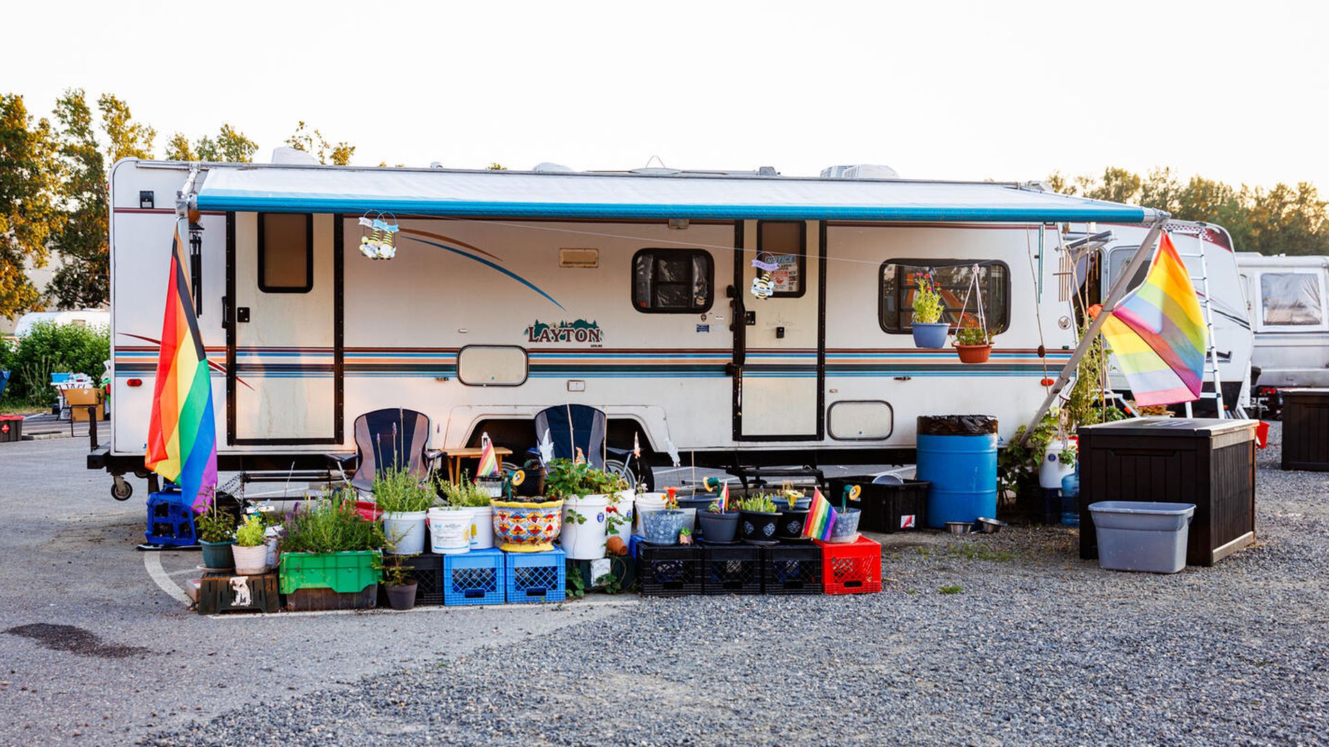 White Layton RV with blue accents, blue awning extended, two rainbow pride flags on each side, black and blue chairs, and colorful potted plants in buckets and crates outside on gravel.