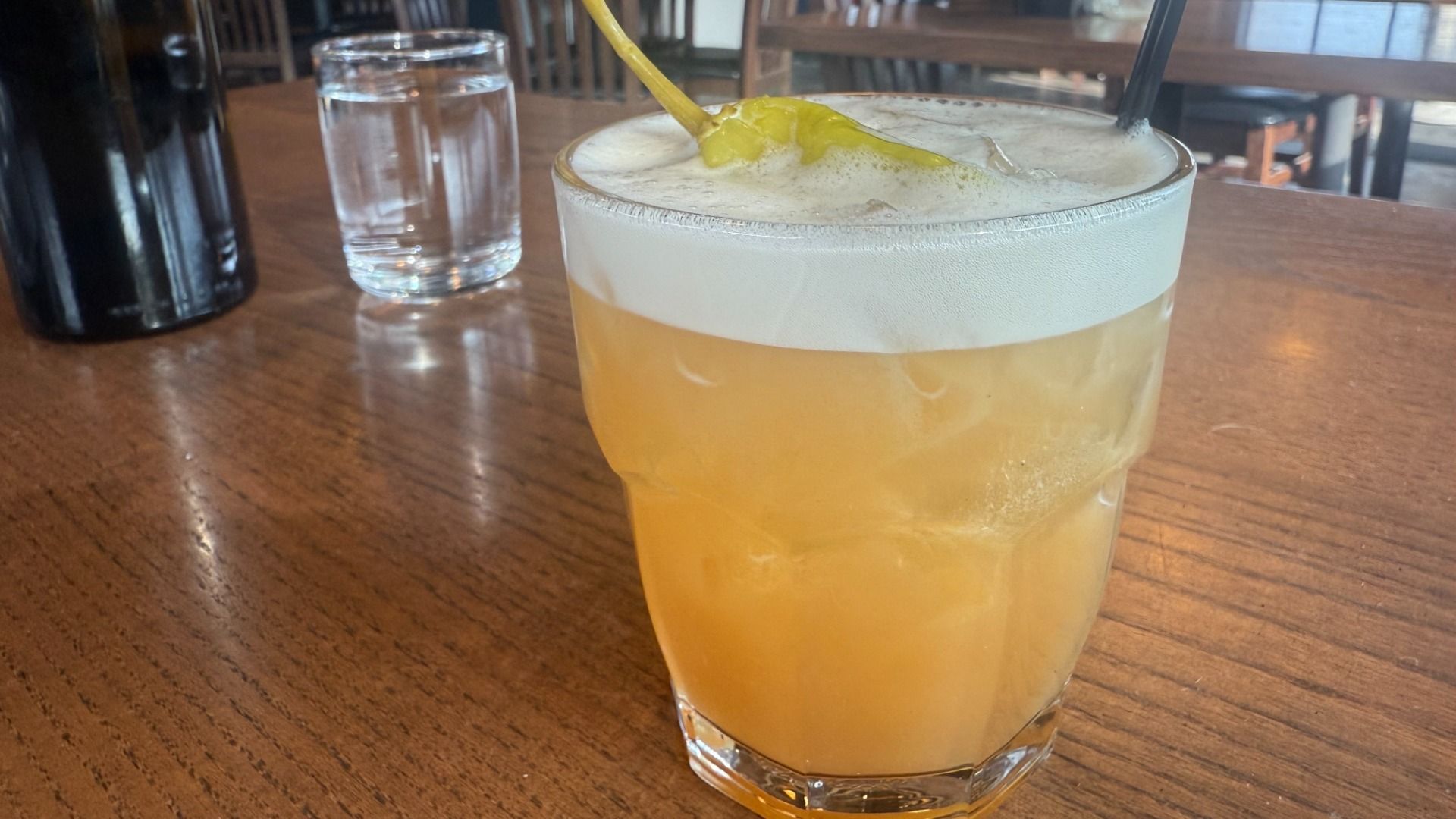 Orange-yellow frothy cocktail in a glass with ice, garnished with a green pepper on a wooden table, with a glass of water and a dark bottle in the background in a restaurant setting.