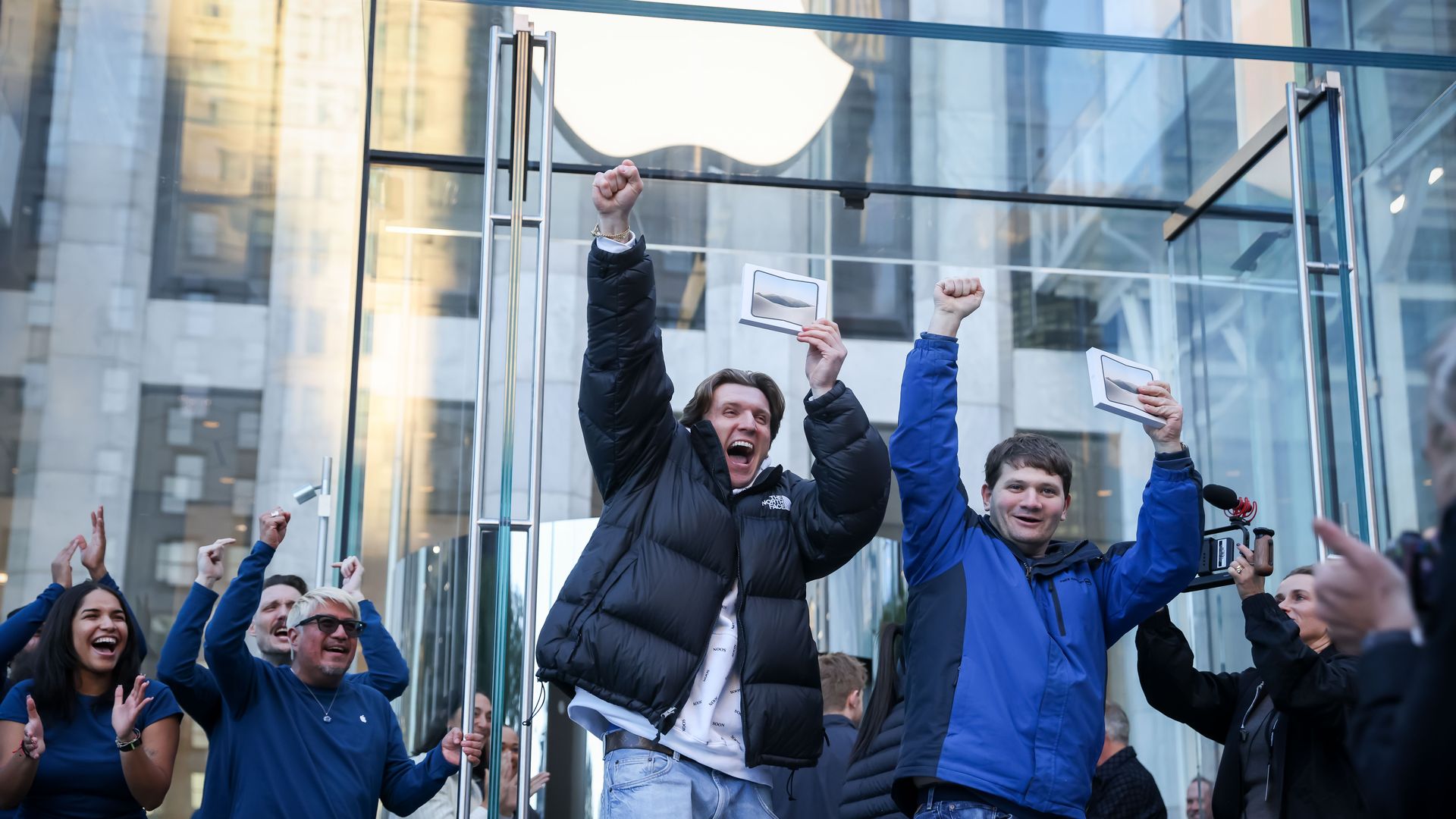 Group of excited people outside a glass storefront with an Apple logo, raising fists in celebration and waving small cards; many wear blue jackets in an urban setting.