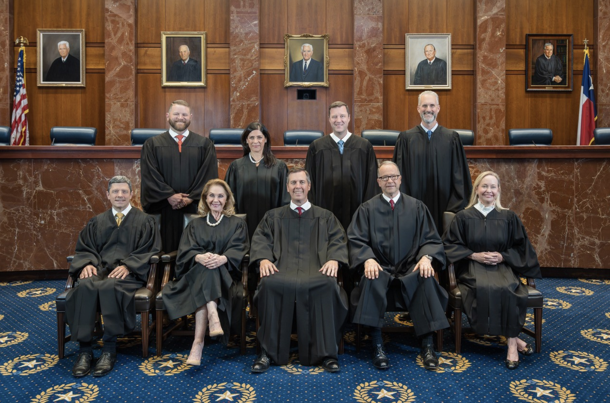 Group of people in black graduation robes posing for a formal photo in a courtroom. They stand and sit in front of a wood-paneled wall with framed portraits and flags on both sides.