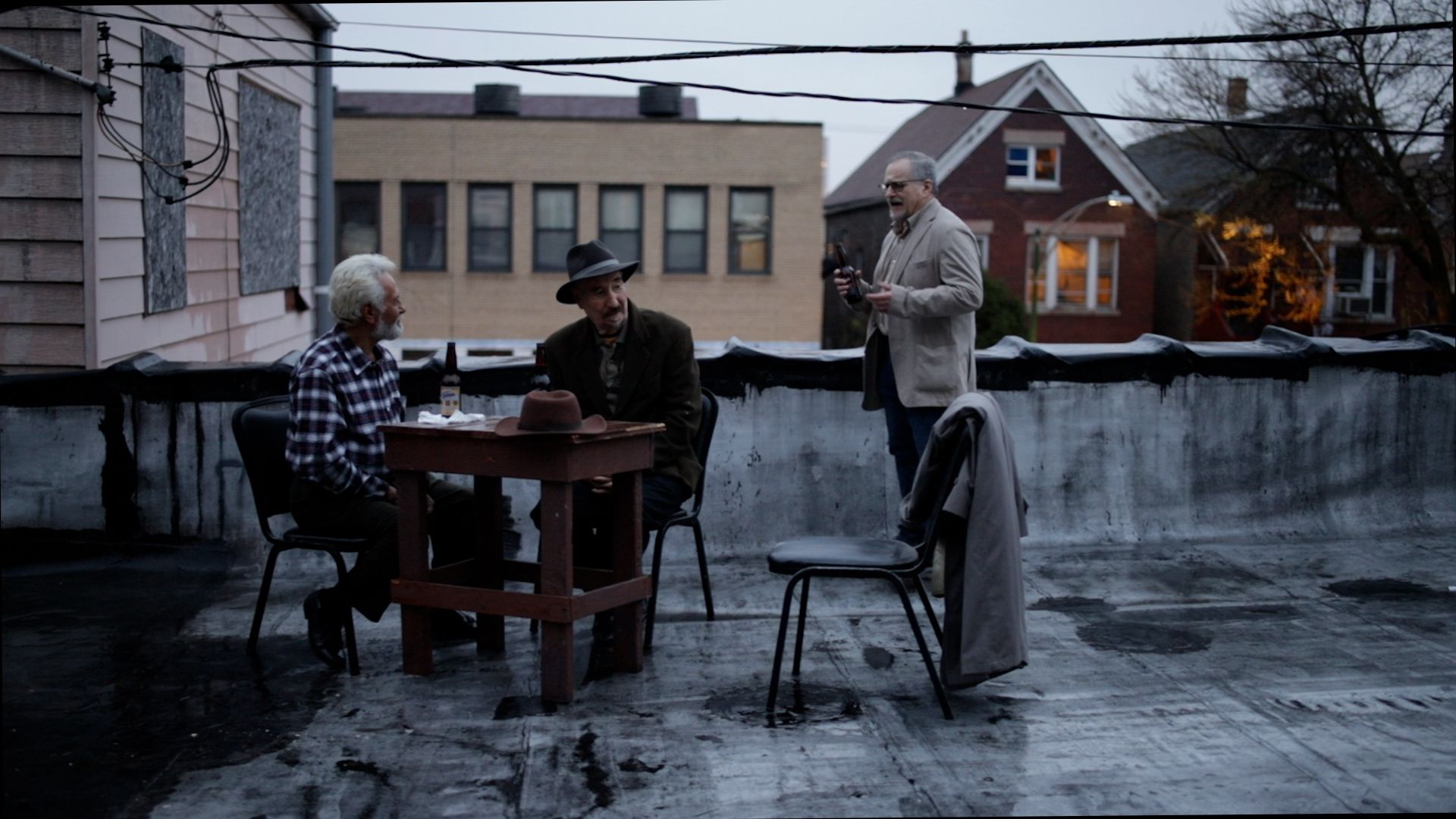 Production still of three actors sitting at a table on a roof.