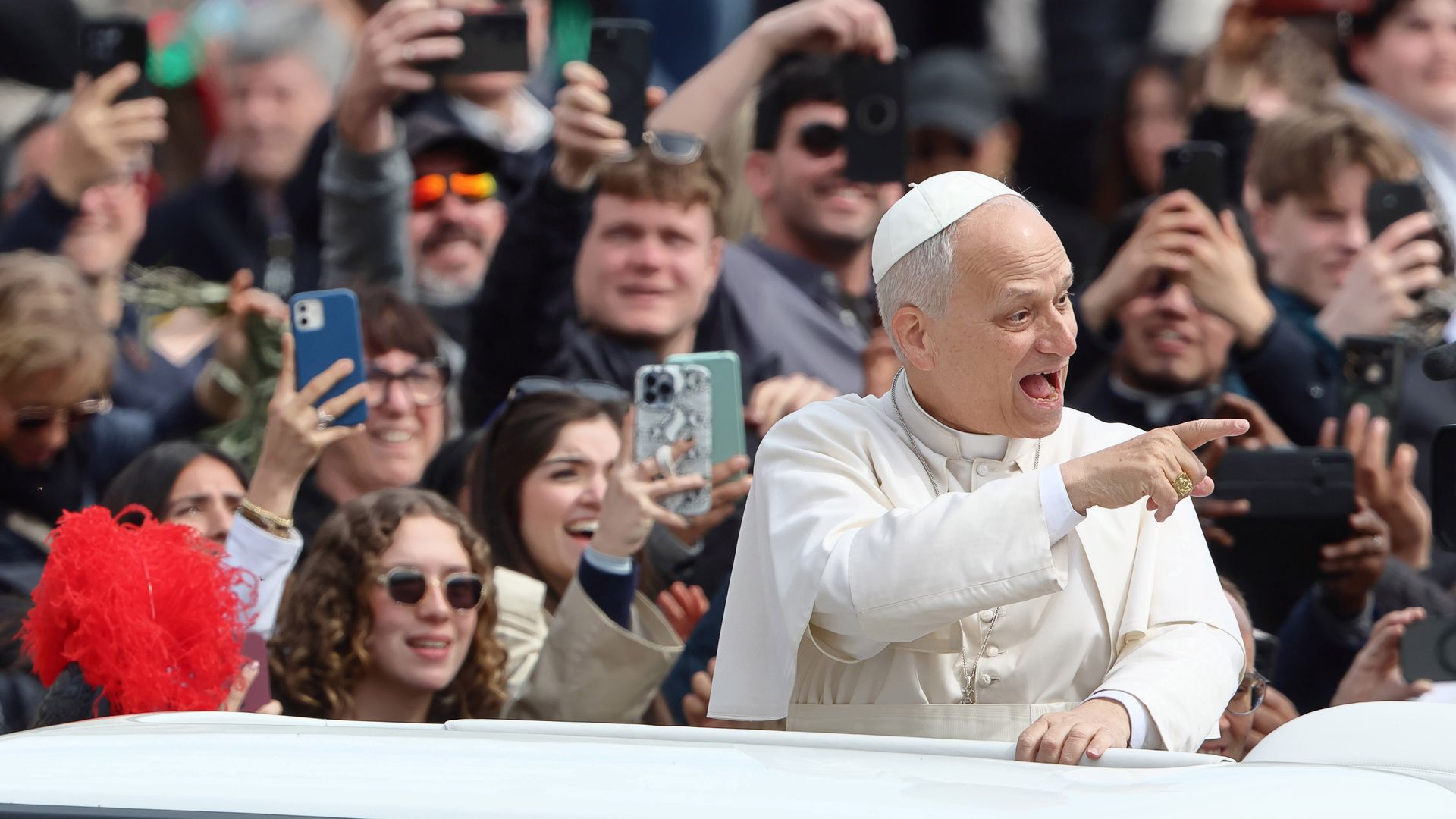 Pope Leo in white papal attire leans from a popemobile, smiling and pointing to the right, as a large crowd of fans behind him take photos with smartphones and cheer.