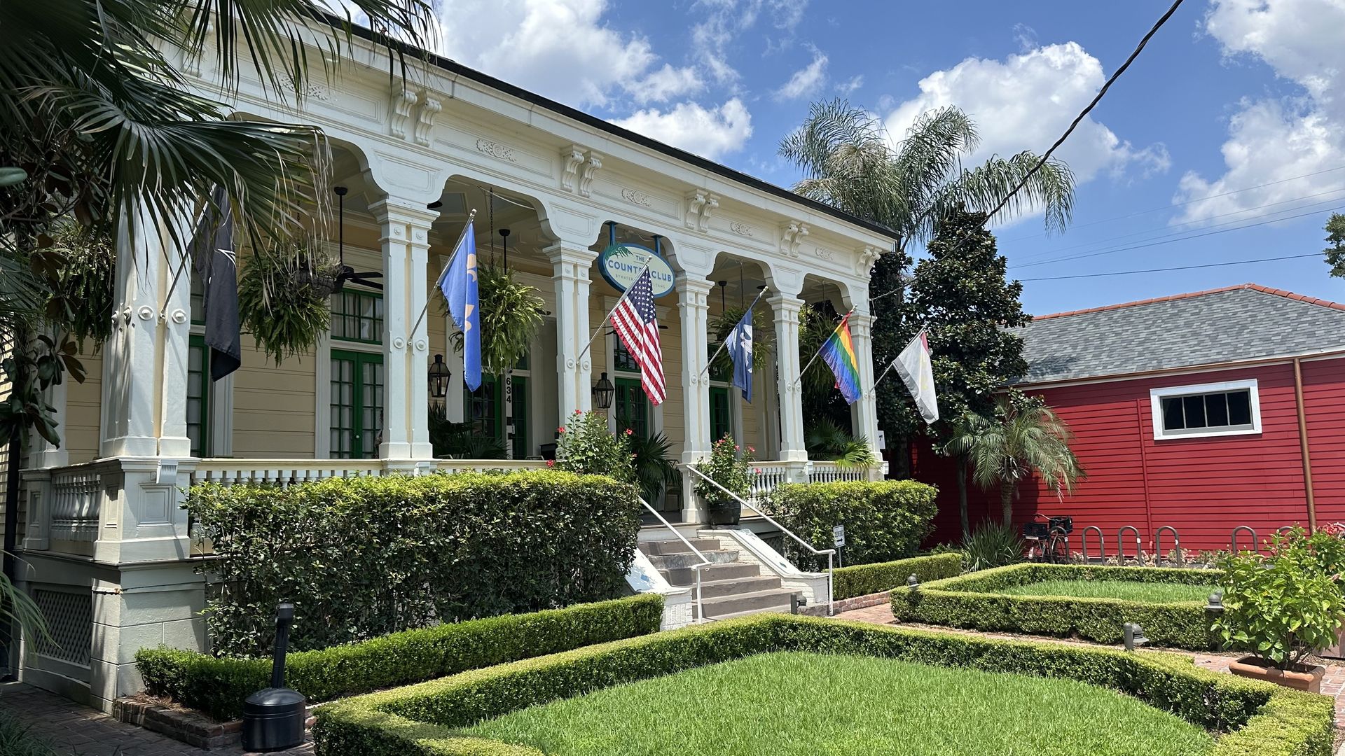 An Italianate cottage has multiple flags hanging out front on a sunny day.